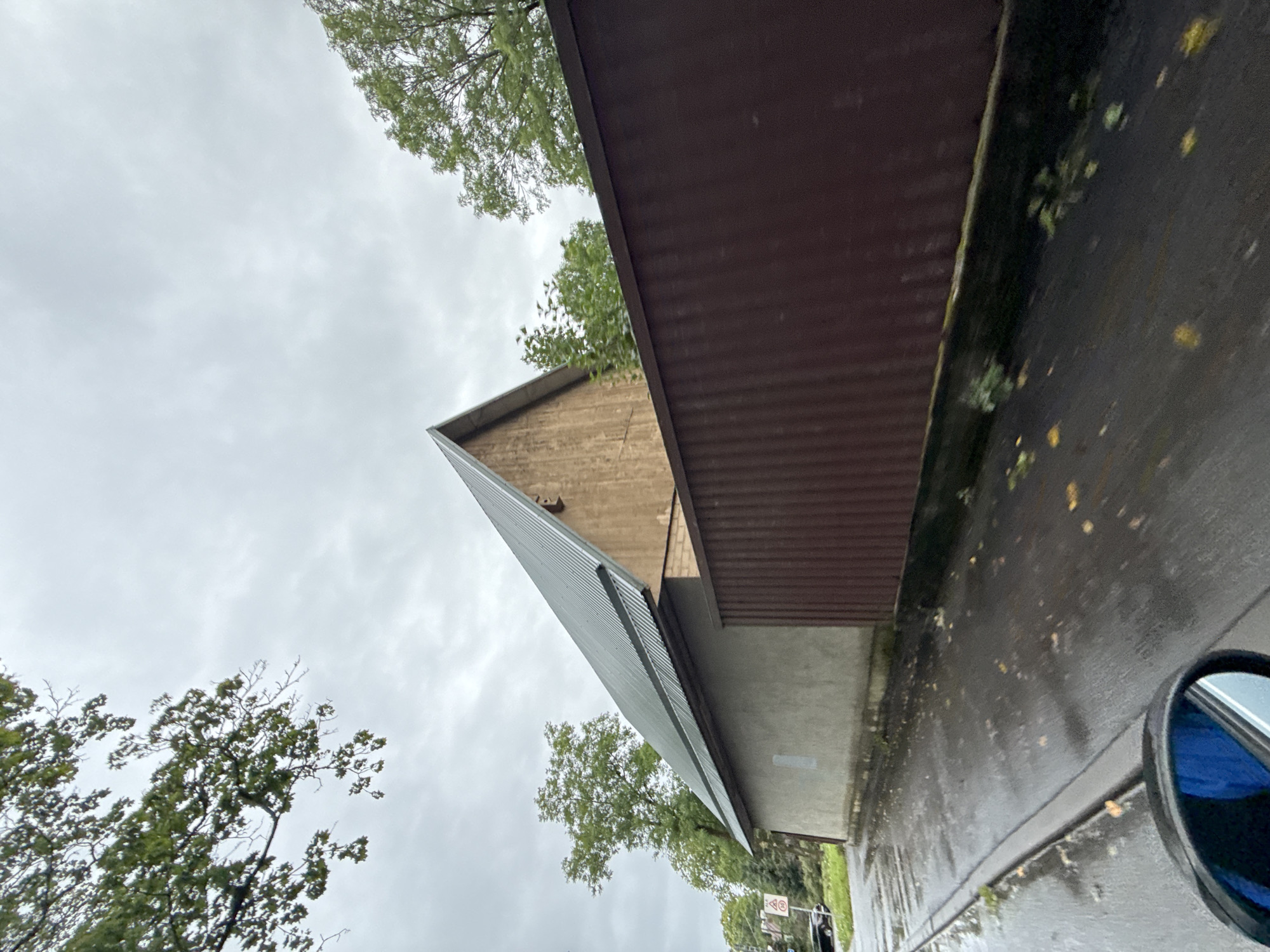 The image depicts a building with a light gray concrete lower section and a wooden upper section with a metal roof. A dark reddish-brown corrugated metal fence runs along the right side of the building, partially obscuring it. The ground in front of the building is wet and reflective, suggesting recent rain. Fallen leaves are scattered on the wet pavement, indicating an autumn setting. Trees with green foliage are visible behind the building and fence, and in the background. A street with a curb is in the foreground, and a car's side mirror, reflecting a blue object, is partially visible in the lower left corner, indicating the photo was taken from a vehicle. A traffic sign is visible on the left side of the street, with a "30" speed limit symbol. The sky is overcast with gray clouds. The scene appears to be in a residential or commercial area.