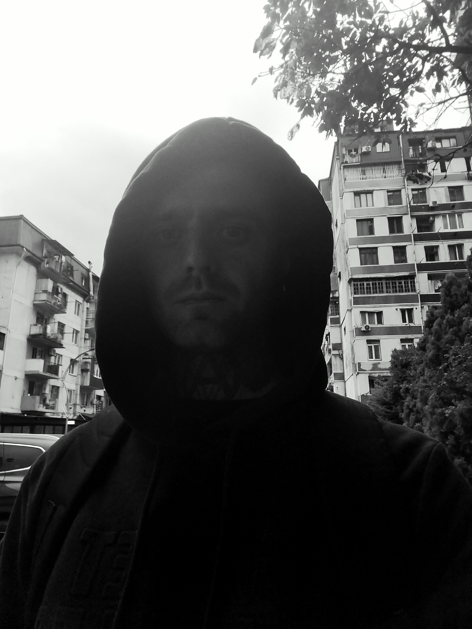 The image is a black and white, high-angle, selfie-style photograph taken outdoors in Batumi, Georgia. The foreground is dominated by the hooded face of a person, with their face mostly in shadow, revealing only their eyes, nose, and mouth. Visible tattoos on the neck suggest a distinctive marking. The subject is wearing a dark hooded garment, obscuring much of their upper body.
Behind the subject, a multi-story residential building with numerous balconies and windows is visible, characteristic of Soviet-era architecture often found in Batumi. Trees and foliage are present to the right of the building. To the left, a vehicle, possibly a car, is partially visible. The overall environment suggests an urban setting, likely a residential street or courtyard. The sky appears overcast, contributing to the muted tones of the black and white image.