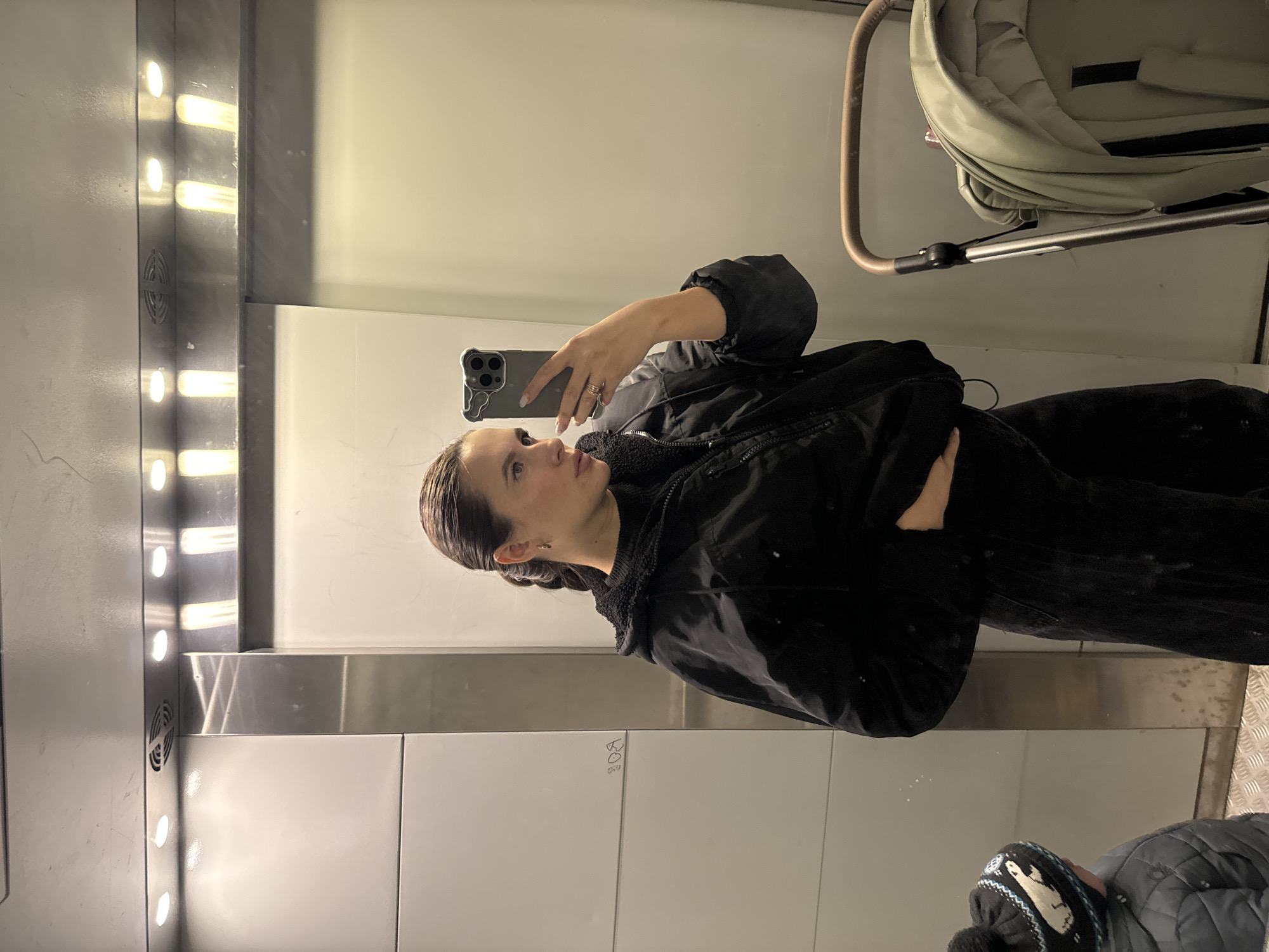 A woman is taking a selfie in what appears to be an elevator or small service room, possibly within a public building. The location cannot be determined to be Odintsovo, Russia with only the visual evidence. The woman is wearing a black jacket and dark pants. She is holding a smartphone with a triple-lens camera in her right hand, presumably to take the picture. The elevator walls are paneled in white, and a strip of vertical lights runs along one side. A baby stroller is visible behind her. Some writing or graffiti can be seen faintly on the wall. A hat and a jacket can be seen on the floor of the elevator.