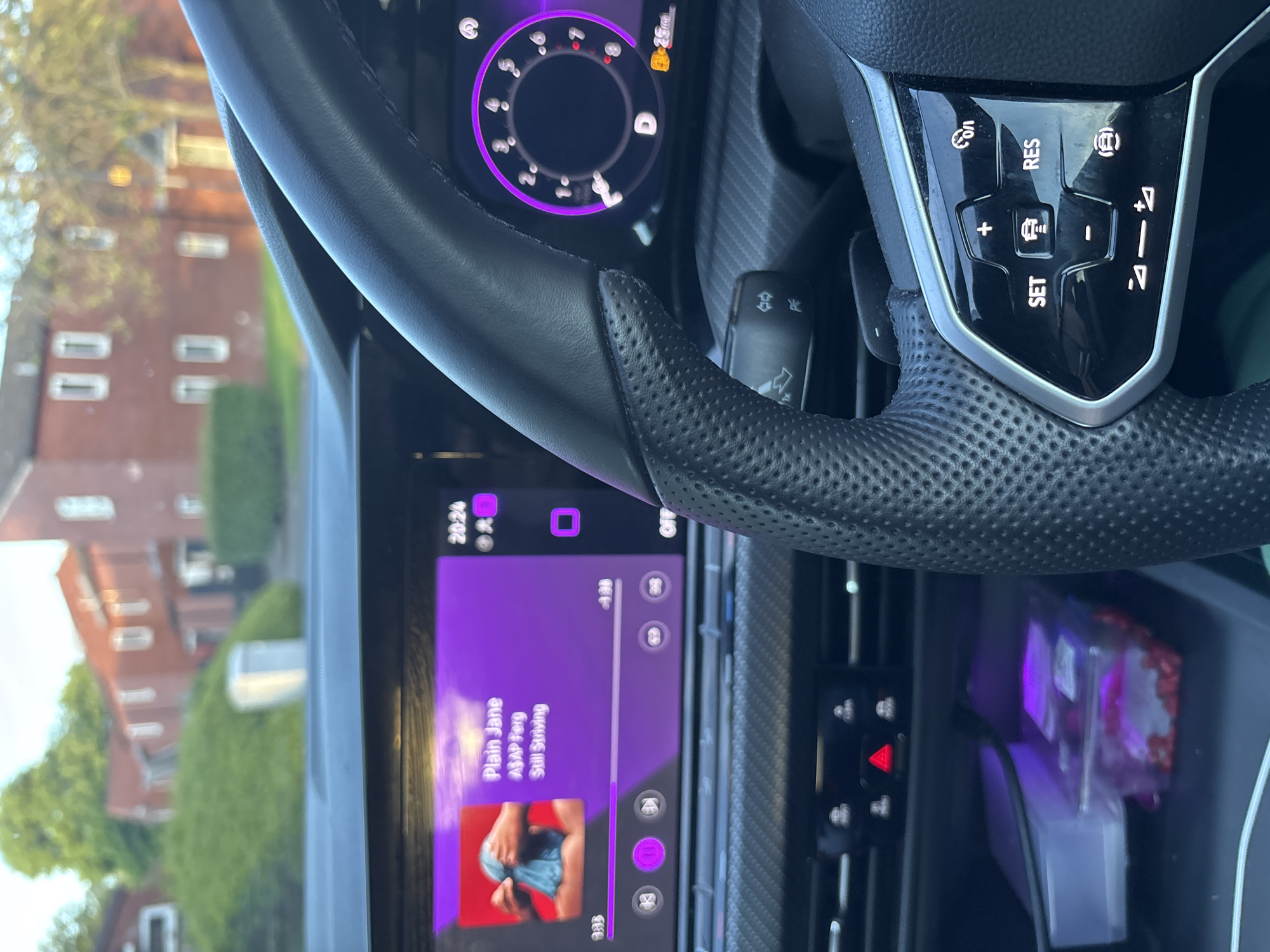 The image shows the interior of a car, specifically the driver's side, with a focus on the steering wheel, dashboard, and infotainment system. The infotainment screen displays the song "Plain Jane" by ASAP Ferg from the album "Still Striving." The digital instrument cluster behind the steering wheel shows the car is in "D" drive mode. The steering wheel has cruise control buttons. A residential area with brick buildings and trees is visible through the windshield, suggesting the car is parked or driving in a suburban neighborhood. A box is visible in the center console of the car.