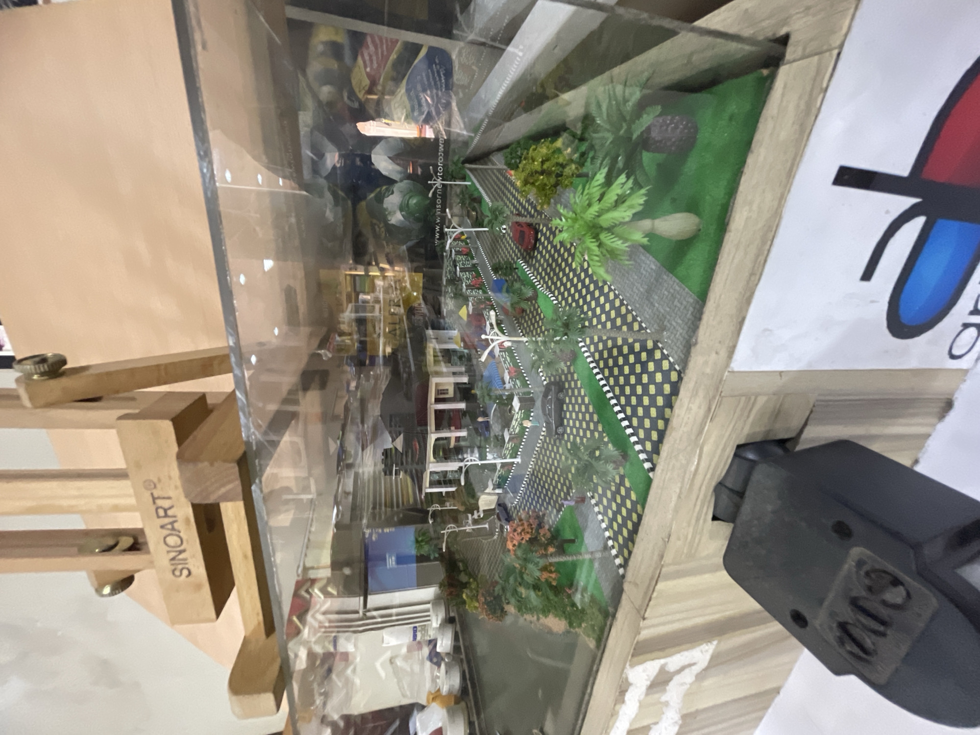 A diorama model of a street scene in Lagos, Nigeria, is displayed within a glass case. The model features miniature buildings, roads with yellow and black checkered edges, trees, and other landscape elements. The diorama is set on a wooden base. The word "SINOART" is visible on a wooden easel supporting the diorama. Part of a logo with red and blue colors is visible on a piece of paper next to the diorama. A black object, possibly a camera or lighting fixture, is attached to the side of the display case. Reflections within the glass show additional items and shelves in the background.