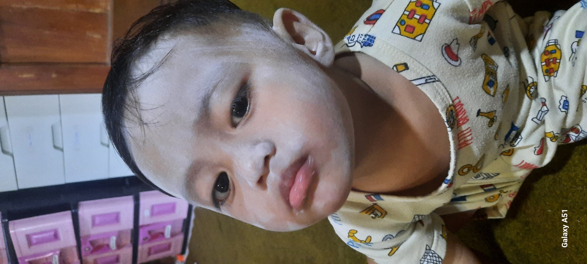A close-up shot shows a young child's face, appearing to be in Jatitengah, Indonesia. The child has fair skin and is wearing a light-colored shirt with a repeating pattern of cartoon characters. The child's face is dusted with a white substance, possibly powder, which is particularly noticeable on the forehead and cheeks. The child's lips are slightly parted, and their expression could be interpreted as pouty. In the background, a multi-tiered pink storage unit is visible to the left, and a wooden cabinet is on the right. The floor appears to be covered in a brown carpet or rug. The bottom left corner of the image displays "Galaxy A51," indicating the device used to capture the photo.