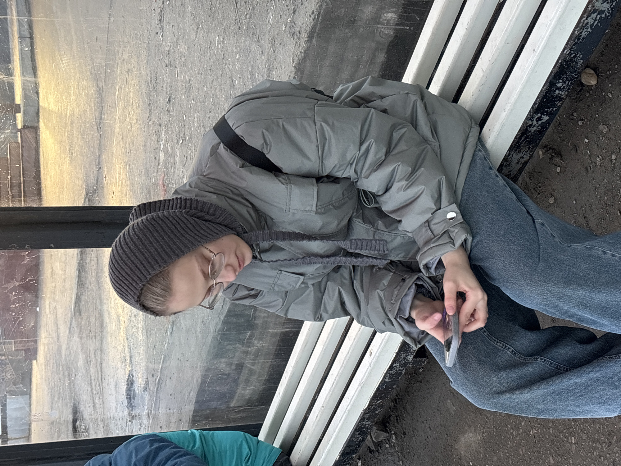 A person wearing a grey beanie, glasses, a grey puffer jacket, and blue jeans is seated on a white slatted bench at what appears to be a bus stop in Perm, Russia. The person is holding a smartphone. The bus stop has a glass panel, and the foreground shows a portion of another person in a blue and green jacket. The background suggests an urban environment.
