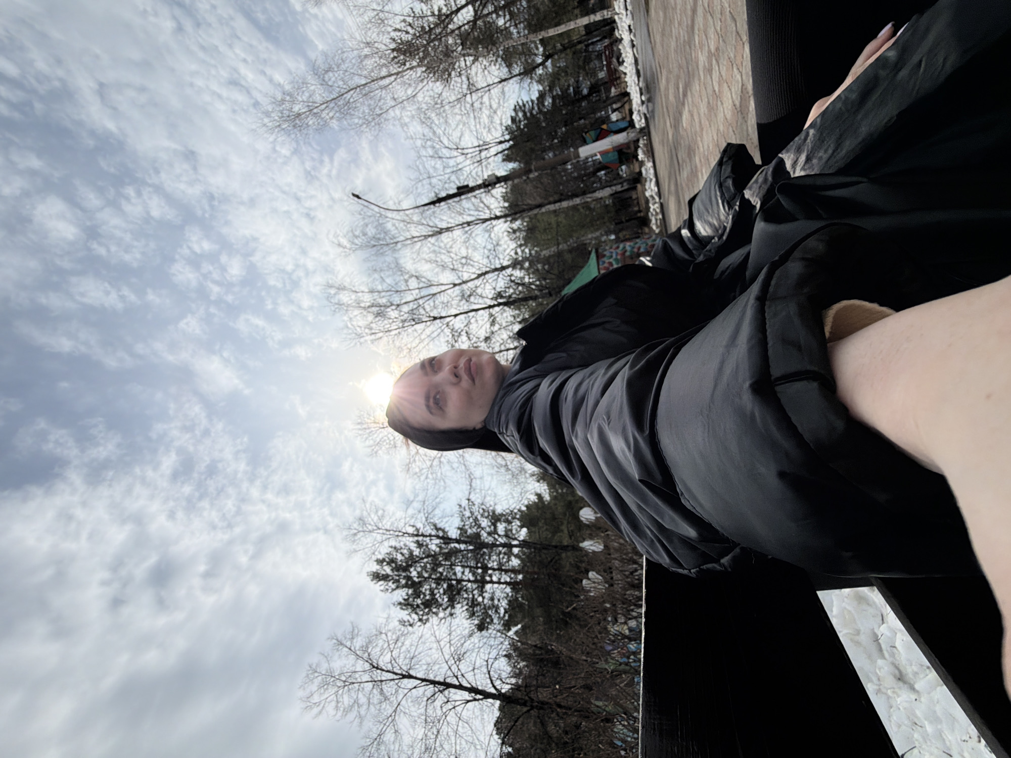A person is sitting on a bench in an outdoor setting in Chaikovsky, Russia. The person is wearing a black jacket. The sky is visible with scattered clouds. Trees are in the background. The sun is shining brightly, positioned near the person's head.