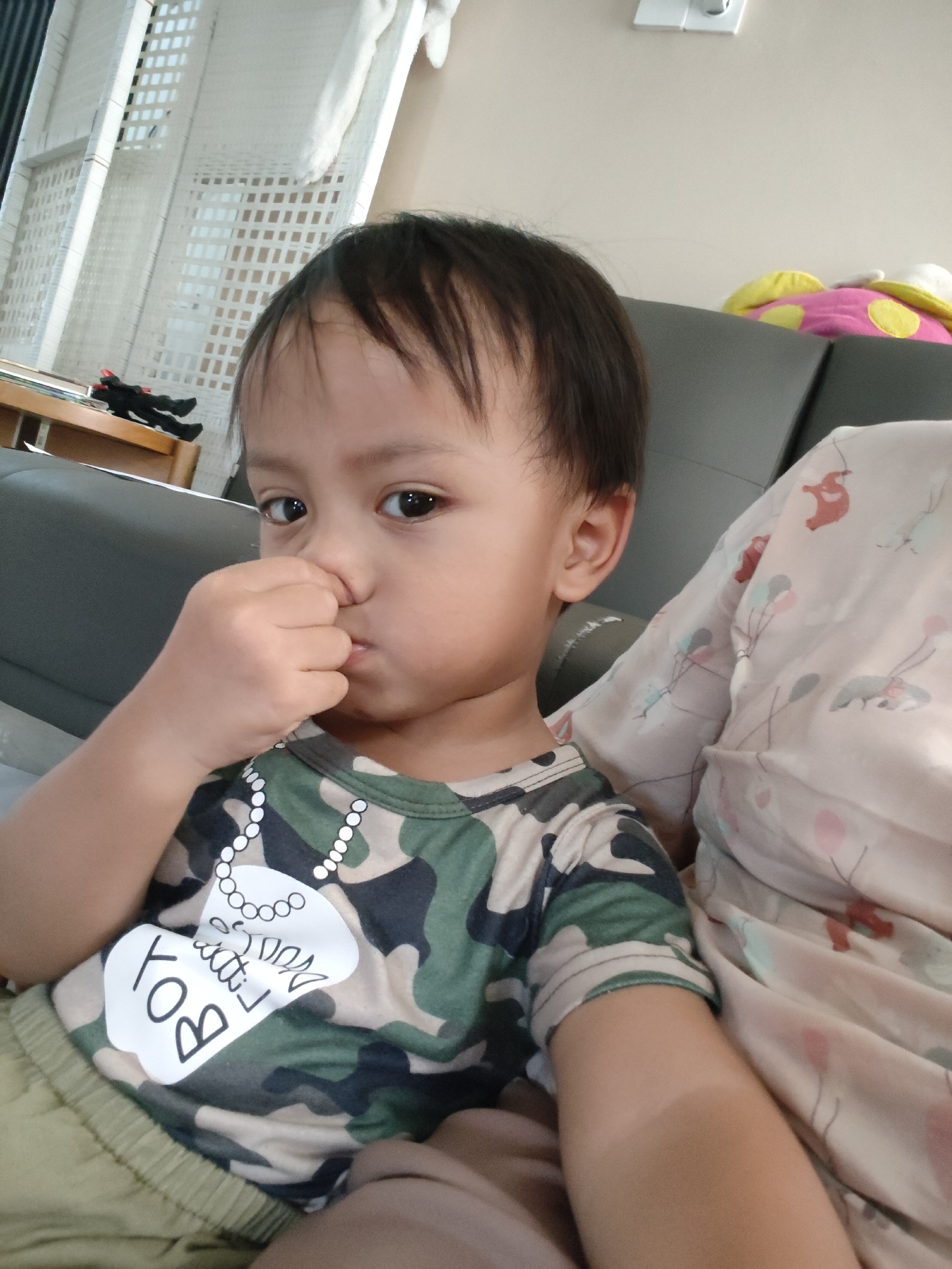 A toddler with dark hair and brown eyes is being held in a seated position. The toddler is wearing a camouflage t-shirt with a white graphic of a dog tag and the word "BOY" on it. The toddler's right hand is touching their nose and mouth. The background consists of a gray sofa, a white woven shelf, and a wall with a light-colored outlet. The overall scene appears to be indoors, likely in a residential setting. There is no visual information in the image that would allow for the determination of its location in Pemalang, Indonesia.