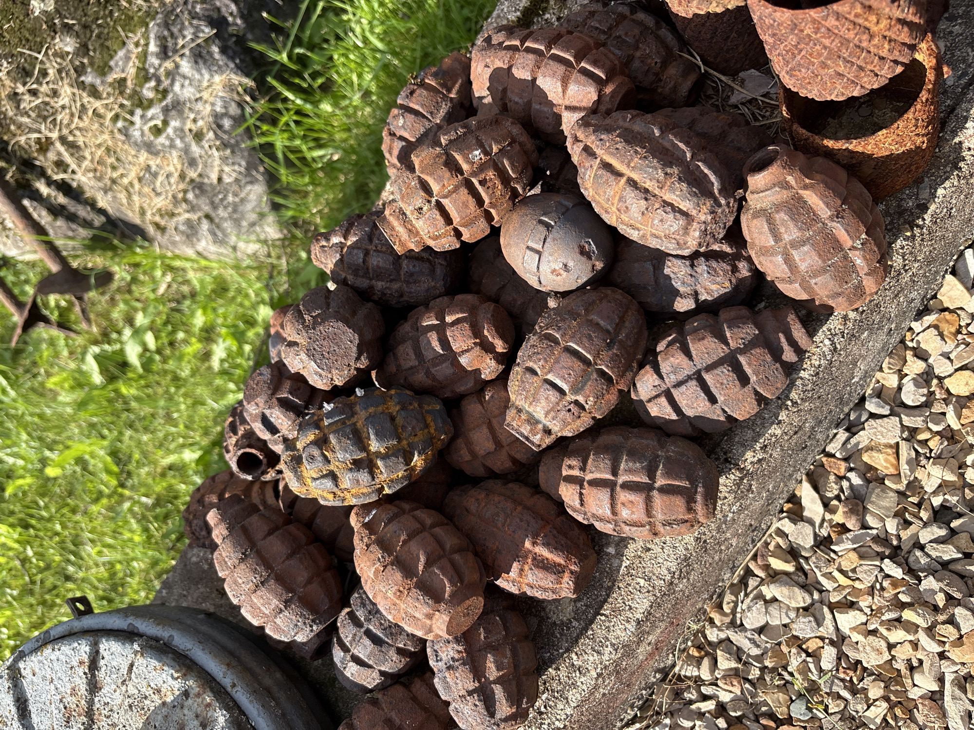 A pile of rusted, cast-iron, pineapple-shaped hand grenades lies on a stone ledge covered in small gravel, with grass and a large rock in the background. One grenade is missing its fuse. This scene is located in the city of Zhalybnya, Russia. There are no people or animals present in the image. The lighting suggests it is daytime.