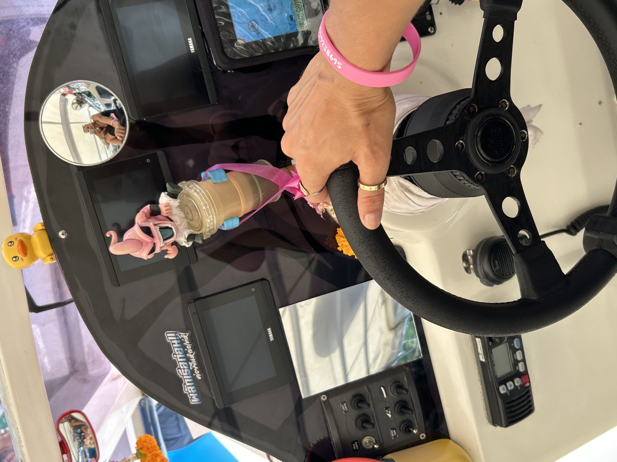 The image depicts the interior of a boat's helm in Ao Nang, Thailand, focusing on a person's hand gripping the steering wheel. The hand is adorned with two rings and a bright pink silicone bracelet with white lettering. On the dashboard, a small yellow duck figurine is positioned near a rearview mirror reflecting a person sitting on the boat. A figurine of Majin Buu from Dragon Ball Z is prominently displayed, holding a cup adorned with pink ribbons. The dashboard also features two Yamaha screens, a control panel with various switches, and a marine radio. The overall environment suggests a recreational boating activity.