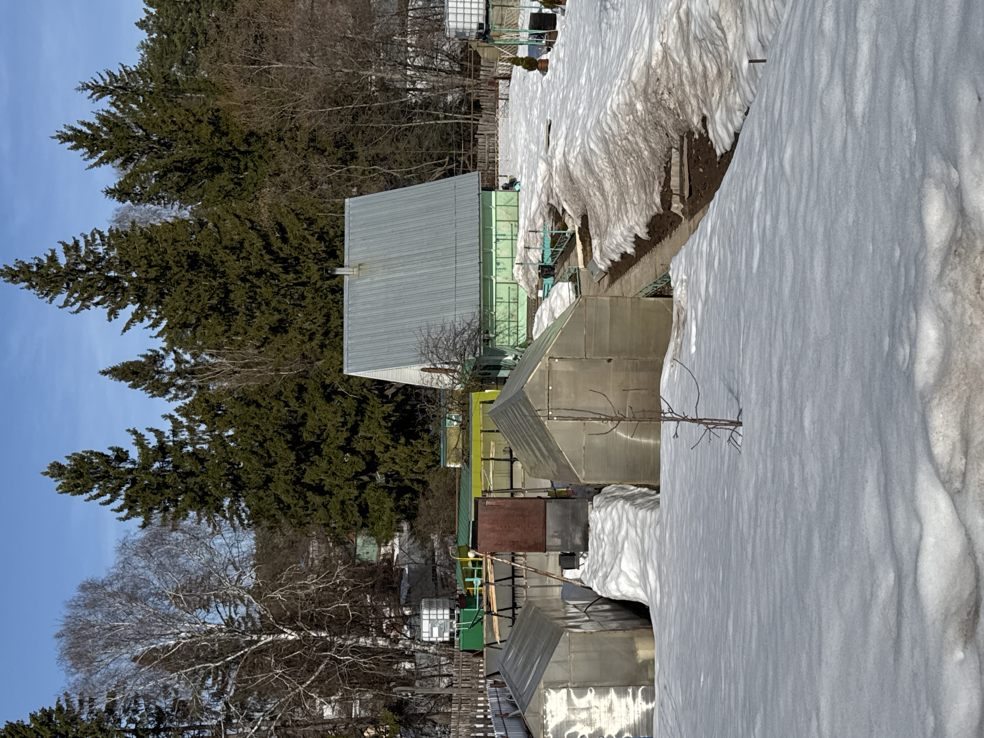 The image depicts an outdoor scene in Chaikovsky, Russia, showcasing remnants of winter transitioning into spring. Predominantly, there are several metallic structures or small buildings, potentially greenhouses or storage units, arranged in a line or clustered together. Heavy snowdrifts are visible, partially covering or abutting these structures, indicating recent snowfall.
In the background, a mix of evergreen and deciduous trees is observed, some still bare while others begin to show signs of foliage. A clear blue sky is visible above the trees, suggesting a sunny day. A fence is also seen along with other indistinct buildings, suggesting this may be a residential or commercial area.