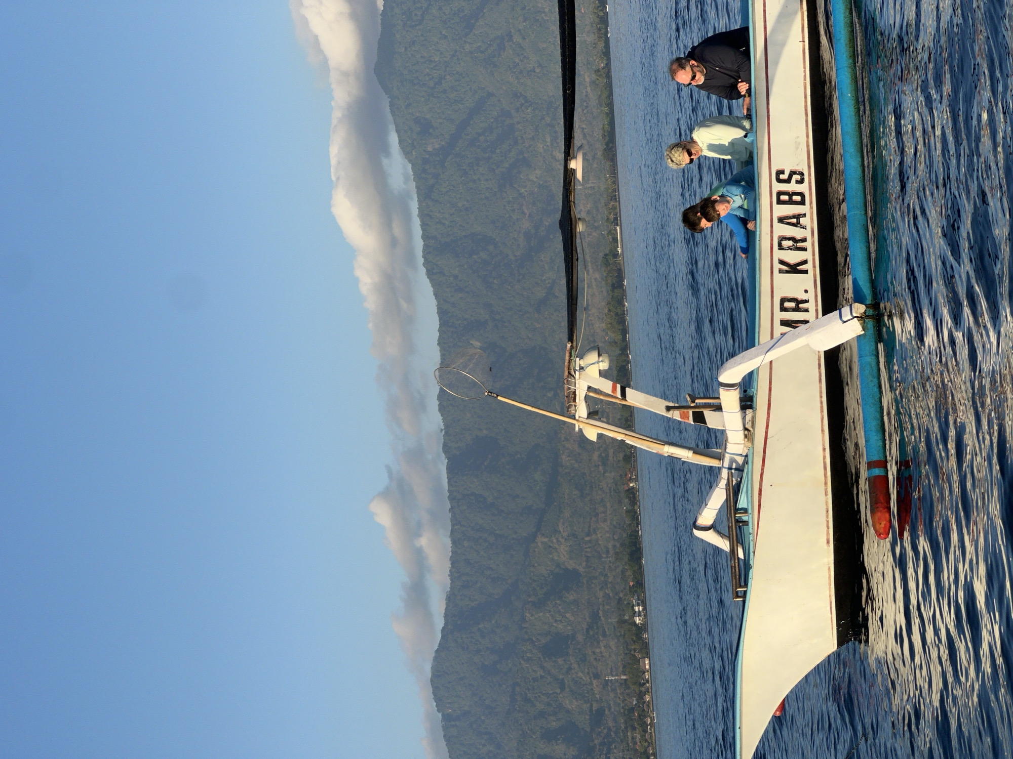Four people are on a boat named "MR. KRABS" on the water. A man in a dark jacket is on the right, a woman in a light-colored hooded sweatshirt is next to him. A young boy in a blue shirt is in front of them, and another person in a blue shirt is behind him. The boat is white with red trim and has a blue float attached to the side. In the background, there is a large, green, tree-covered mountain range with a town at its base. The sky is blue with some white clouds. The water is choppy.