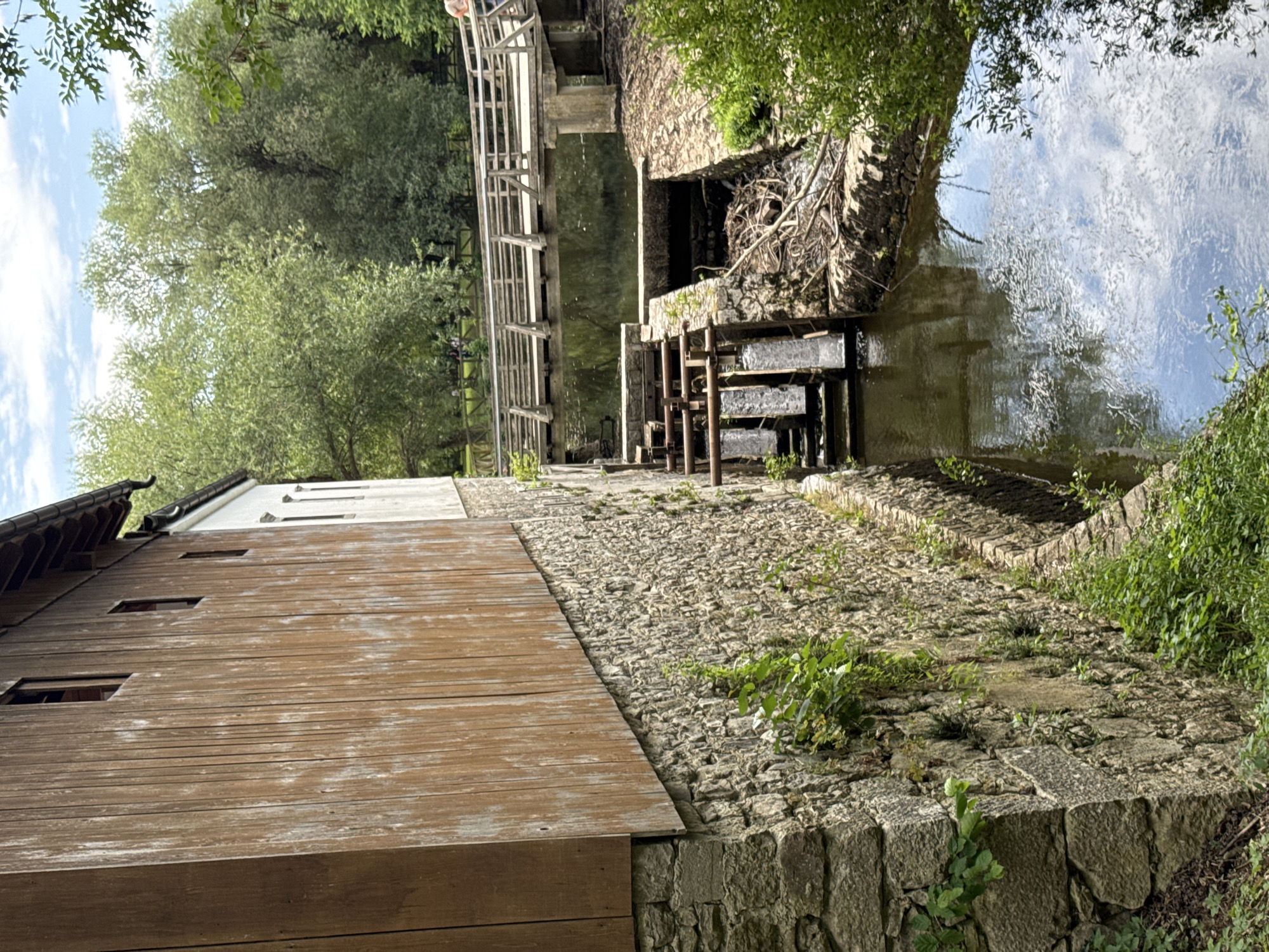 The image shows a historic watermill structure situated on the edge of a flowing body of water, likely a river or stream, in Postojna, Slovenia. The mill building itself is constructed with a stone foundation and a wooden upper story featuring weathered planks and small rectangular windows. Adjacent to the mill, a water wheel mechanism with wooden paddles is visible, indicating its operational state. The water flows through a channel formed by stone walls, with debris and branches accumulated in one section. A wooden bridge spans the water further downstream, with a few individuals faintly visible on it. Lush green trees and vegetation surround the watercourse and the mill, suggesting a natural, park-like setting. The sky above is partly cloudy. The scene captures a rustic and functional architectural element integrated within a verdant landscape.