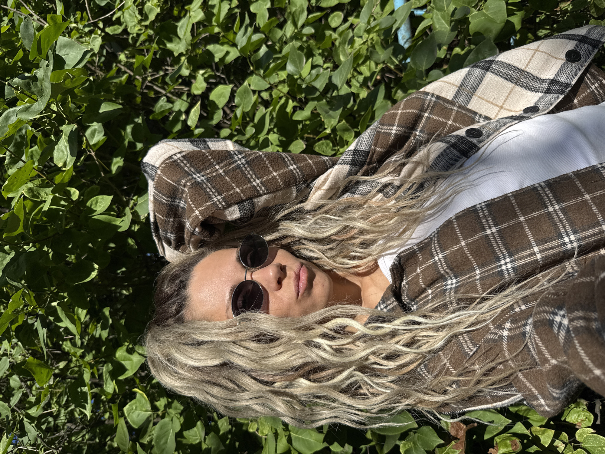 A woman is captured in a selfie taken outdoors against a backdrop of lush green foliage. She has long, blonde, wavy hair tied up in a high ponytail and is wearing oval-shaped sunglasses. Her attire consists of a white ribbed tank top beneath a brown, beige, and white plaid shirt jacket, which is unbuttoned. The woman is posing with her left arm bent, hand resting on her head, and her head is tilted slightly to her right. The sunlight casts shadows on her face. The background is dominated by the dense green leaves of a bush or tree, with a hint of a blue object, possibly a fence or structure, visible on the far right. The scene appears to be set in a natural environment, such as a park or garden, within the city of Rusanovo, Russia.