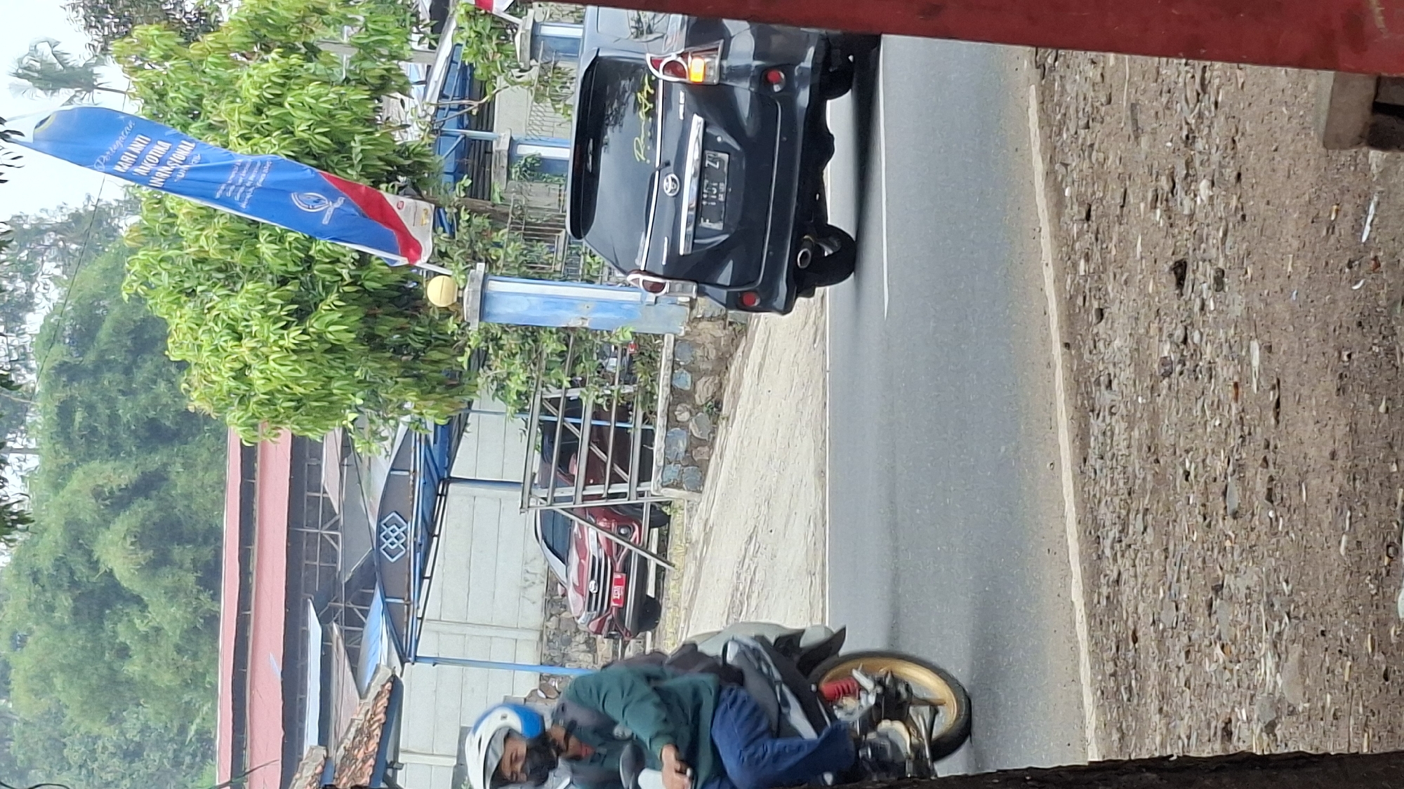 A medium-angle, slightly elevated, full shot captures a street scene in Cilaku, Indonesia, with a focus on vehicles and a blue and red flag. In the foreground, a person on a motorcycle is partially visible from the left, facing right. The rider wears a green jacket, blue jeans, and a white helmet with blue accents, and appears to be looking forward. The motorcycle has a silver body and a gold-colored wheel with red accents.
Further down the road, a dark grey multi-purpose vehicle (MPV) is traveling away from the viewer, with its rear license plate visible. The license plate reads "F 1157 ZY". The MPV has a spoiler and decorative yellow lettering on its rear window. To the left of the MPV, a red SUV is parked behind a metal fence, partially obscured by foliage.
In the background, a building with a red roof is visible, topped by dense green foliage. A tall, blue and red flag, with Indonesian text on it, is prominently displayed to the right of the red SUV and behind the MPV, fluttering in the breeze. The flag has an emblem with a circular design and text. The environment appears to be a road with a paved surface and a rough, gravelly shoulder. A white line marks the edge of the road. The overall lighting suggests an overcast day.