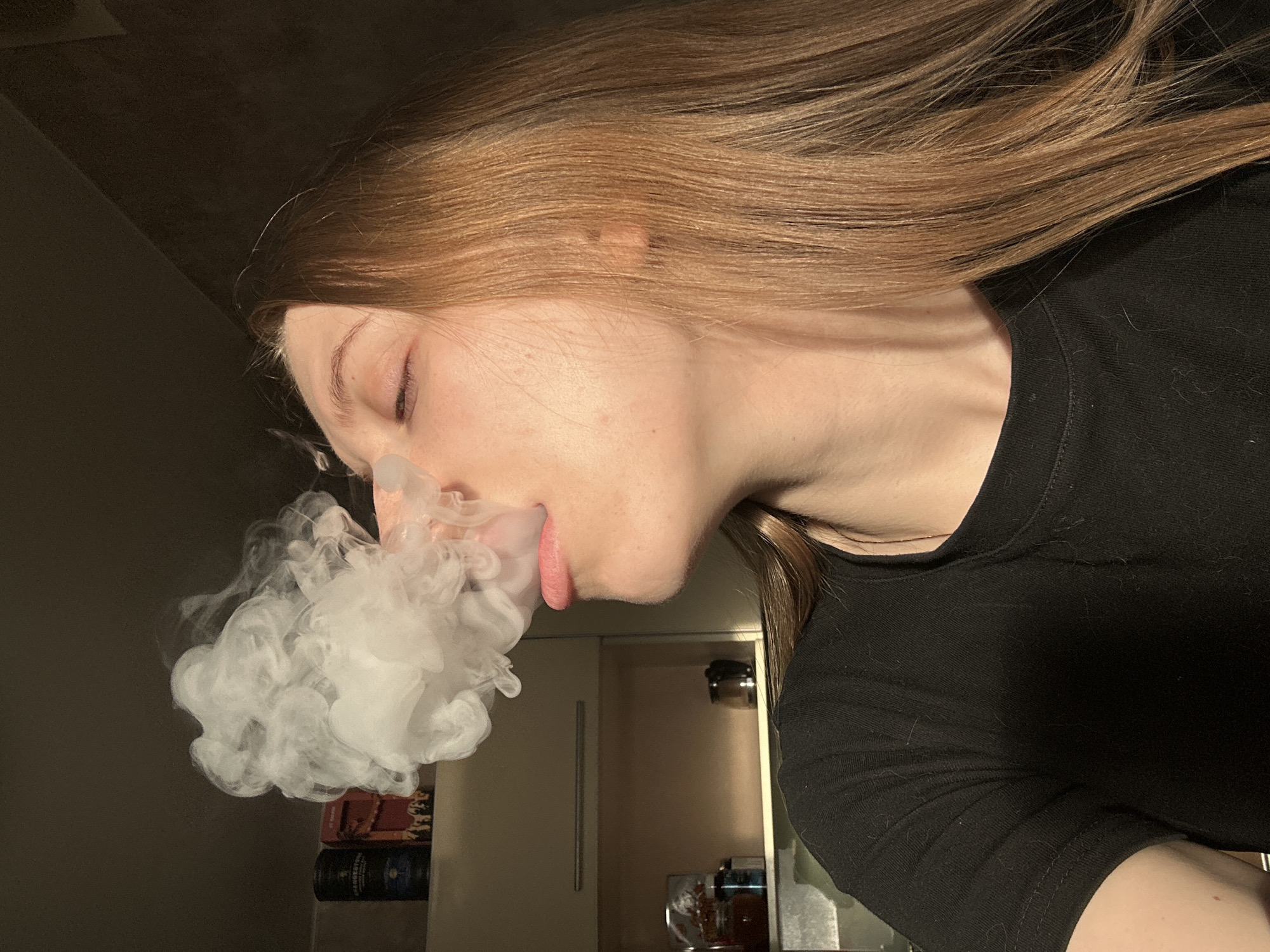 A person with light brown hair and fair skin is shown in profile, exhaling a large cloud of white vapor. The person is wearing a black long-sleeved shirt. The background is slightly blurred, showing a room with a neutral-colored wall on the left, a light-colored cabinet door in the center, and some items on a shelf below the cabinet. The items appear to be a stack of boxed goods, and some kitchenware. The location is not discernable to Chaikovsky, Russia.