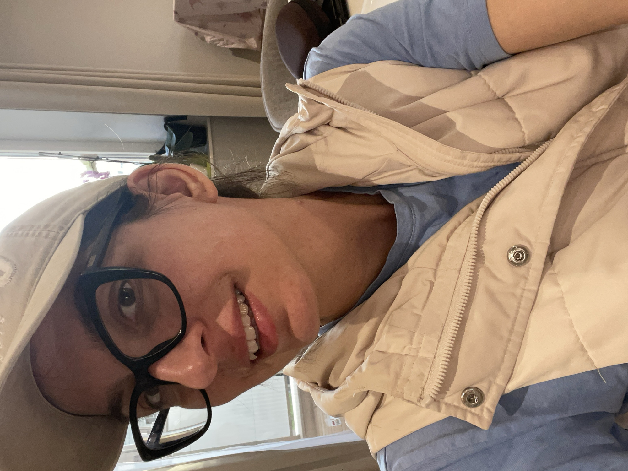 A woman wearing a light-colored baseball cap and large, black-rimmed glasses smiles towards the camera. She is wearing a light blue t-shirt and a light beige, quilted vest with metal snap closures. The background is a room with a window and a curtain or blind. There is no indication of the location being Sofia, Bulgaria.