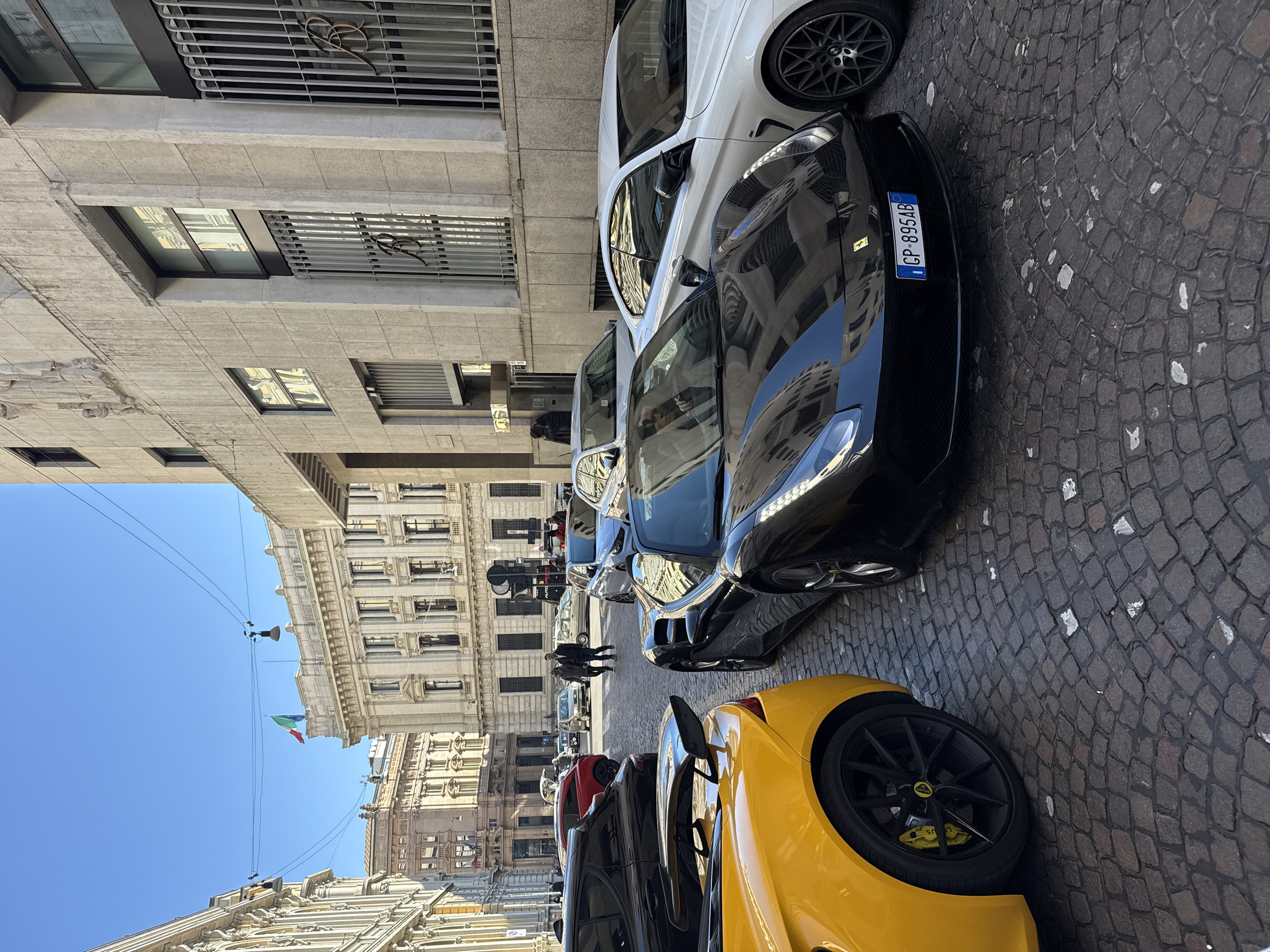 The scene depicts a street in Milan, Italy, specifically in the vicinity of Largo della Scala, characterized by prominent historical architecture. The street surface consists of dark grey and reddish cobblestones with sporadic white debris. The sky is clear and blue, indicating bright daylight conditions.
In the immediate foreground on the left, the rear section of a yellow Lotus sports car is visible, featuring a black multi-spoke wheel with a yellow brake caliper bearing the Lotus logo, and a pronounced black rear spoiler. Adjacent to it, in the center foreground, is a black Ferrari sports car (model likely 458 or similar), parked and facing away from the camera. Its daytime running lights are illuminated, and reflections of the surrounding buildings are visible on its glossy surface. The front license plate, "CP 895AB," is Italian. To the right of the Ferrari, the front section of a white BMW car with a dark multi-spoke wheel is partially visible.
In the midground, several other vehicles are present: a light silver hatchback, a dark sedan, a red hatchback with its trunk open, and a smaller, light-colored sedan in motion further down the street.
Several individuals are present in the midground and background. Two figures walk away on the left sidewalk. Two individuals stand near the red hatchback with its open trunk. Two males walk towards the camera on the right sidewalk. Additional indistinct figures are observed in the background.
The right side of the image is dominated by a large, multi-story, light-colored stone building with a monumental façade. It features several tall windows protected by vertical grates, some of which incorporate a stylized "BR" monogram. Sculpted figures are visible on the building's upper facade. An entrance area, partly recessed, displays a metallic canopy and a prominent "BR" logo. Reflections of other buildings are visible in its windows. This building is identifiable as the former Banca Commerciale Italiana headquarters.
On the left side of the street, multiple ornate, classical-style buildings with intricate facades, columns, and numerous windows line the street. In the background, another multi-story building displays an Italian flag flying from a balcony. Overhead wires traverse the scene, from which streetlights are suspended. Traffic signs are faintly visible in the distance.