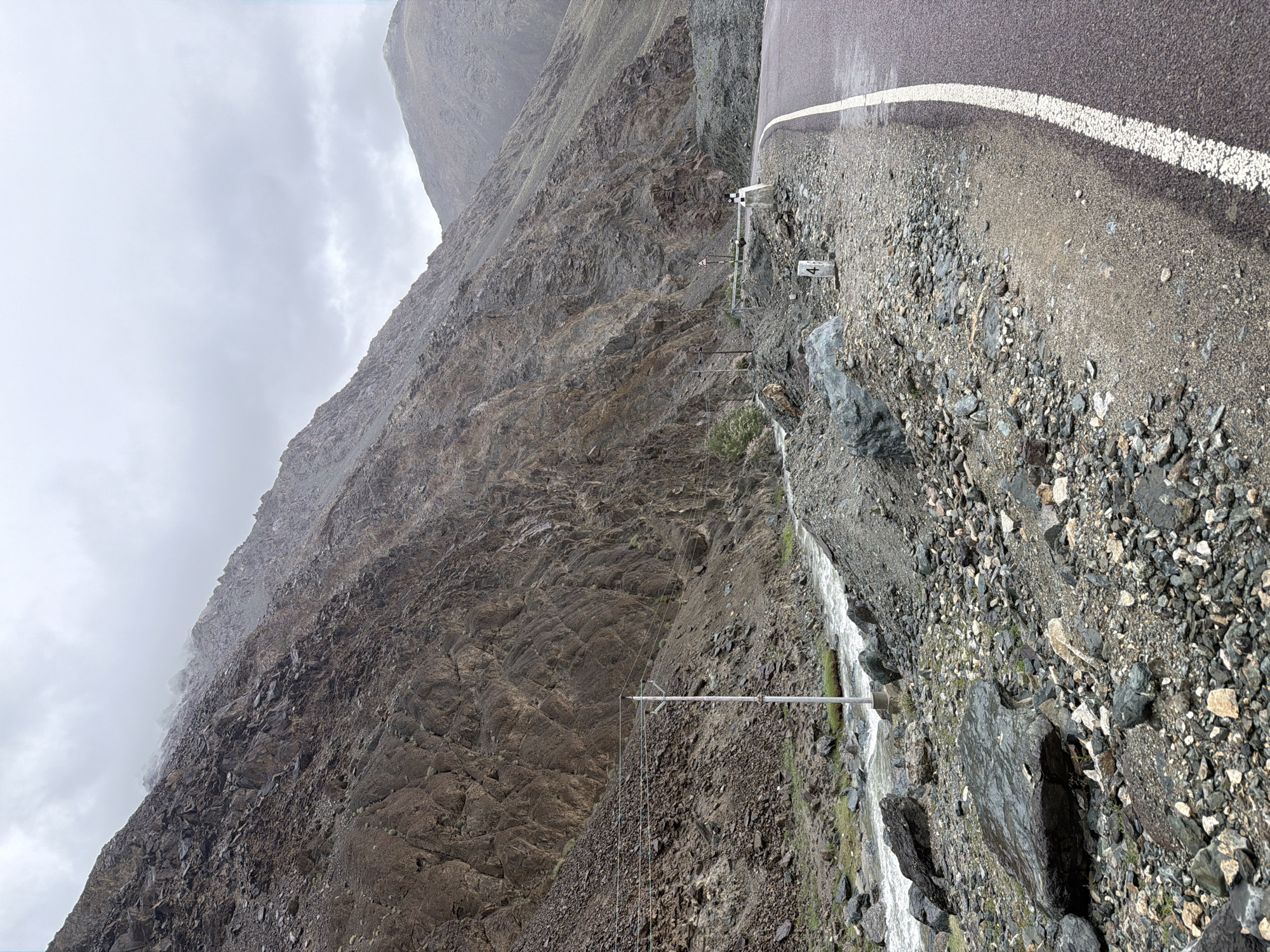 The image depicts a dramatic mountain pass landscape near Sumdo, India, under a cloudy sky. A paved road with a white dividing line curves to the right. To the left of the road, a steep, rocky mountainside rises sharply, showing layers of brown and gray rock formations. A river flows through a rocky ravine at the base of the mountain. Several telephone poles with wires are visible along the mountainside. The ground beside the road is covered in gravel and scattered rocks. A white post with the number "4" marked on it stands near the edge of the road. In the middle ground, a person in white clothing is standing near a guardrail. The overall atmosphere is rugged and remote, with signs of human infrastructure juxtaposed against the raw natural environment.
