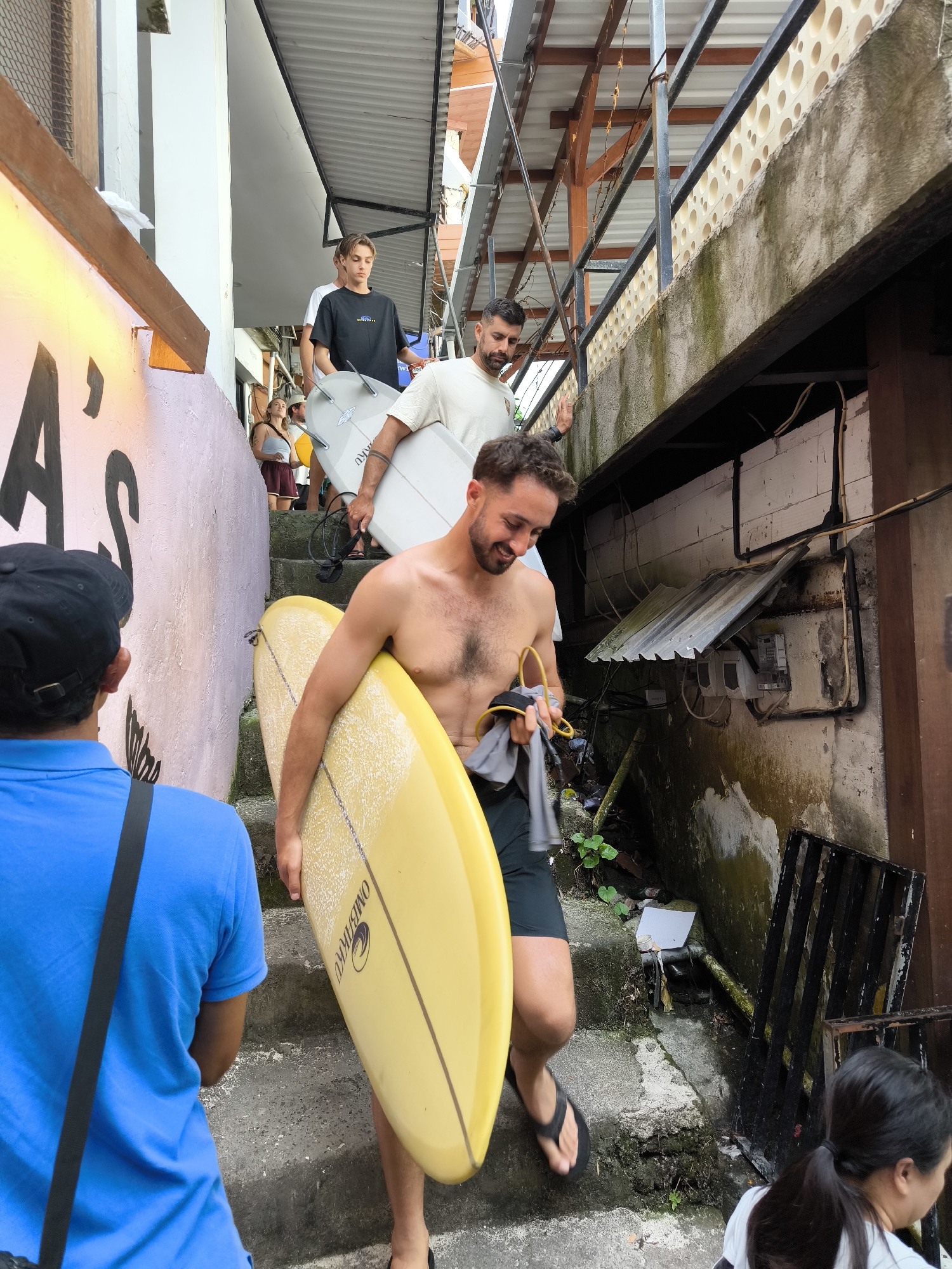 The scene captures a narrow, concrete stairway in Uluwatu, Indonesia. Three men, each carrying a surfboard, are ascending the steps. The man in the foreground is shirtless, holding a yellow surfboard, while the two behind him carry white surfboards. Other people are present, both ascending and descending the stairs. Graffiti adorns the wall to the left, and a combination of concrete, metal, and vegetation makes up the immediate environment surrounding the stairway. Evidence of infrastructure, such as pipes and electrical boxes, is visible on the adjacent wall.