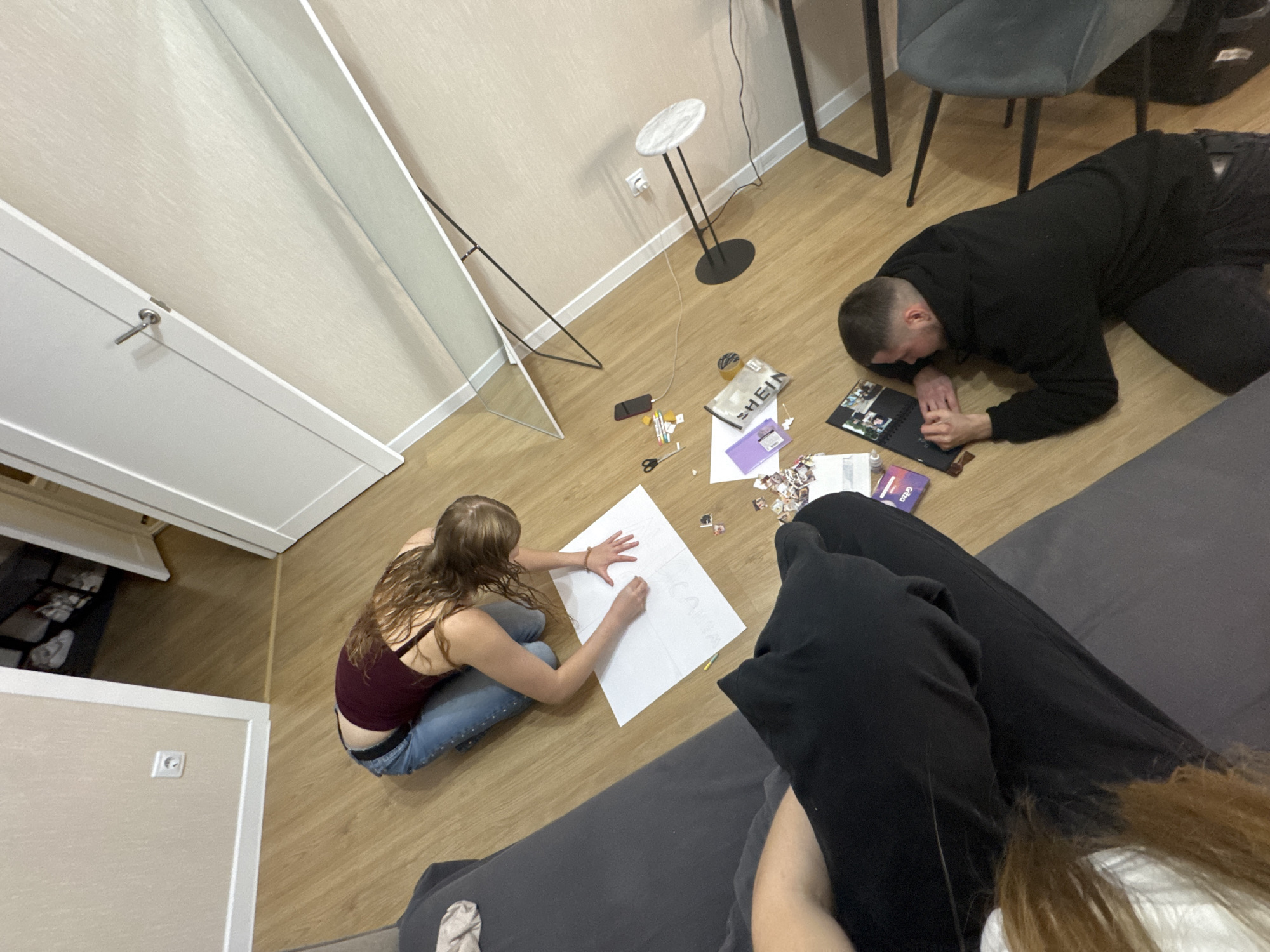 An overhead, wide-angle shot captures two individuals on the floor of a room with wood-like laminate flooring. In the middle ground, a person with long, light brown hair, wearing a maroon tank top and blue jeans, kneels on the floor and appears to be drawing or writing on a large sheet of white paper. Scattered around them are various small items including scissors, pens, and what appear to be printed photographs. To the right, a man wearing a black hooded sweatshirt and dark pants lies on his stomach, looking at an open photo album. In the background, a tall, full-length mirror stands against a light-colored wall. A doorway is visible to the left, showing a sliver of another room. The setting is a domestic interior, consistent with an apartment or house. The location of Perm, Russia, is not discernible from the image itself.