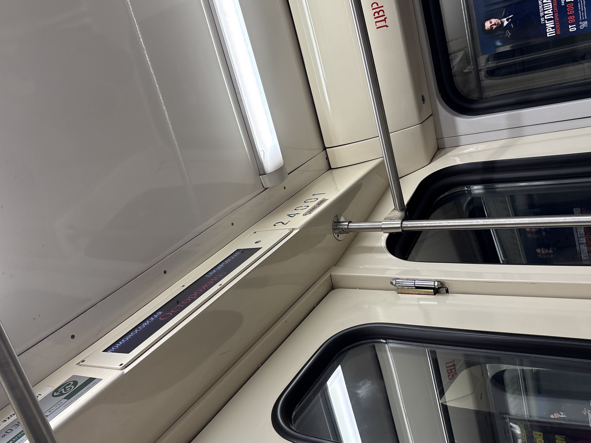 The image captures the interior of a subway train car, likely in Saint Petersburg, Russia. The scene is dominated by the beige and white color scheme of the train's interior walls and ceiling. A long fluorescent light fixture illuminates the left side of the frame, casting a bright glow.
Along the upper wall, above the windows, a digital display is visible, showing Russian text indicating station names: "ЛОМОНОСОВСКАЯ" (Lomonosovskaya) and "ЕЛИЗАРОВСКАЯ" (Yelizarovskaya), which are metro stations in Saint Petersburg. Below these names, the word "Осторожно," (Caution,) is displayed, suggesting the doors are about to close or the train is departing.
Below the display, a small plate reads "81-724," identifying the train car model, and the car number "24001" is also prominently visible. Other text like "320" and "ВАГОН" (Car) are present. Polished metal handrails run horizontally across the top section of the windows, and vertical poles are also visible, providing support for passengers.
Through the train windows, reflections of the opposite side of the car can be seen, including part of an advertisement. This ad, identified by its text, is from "ГУП 'ПЕТЕРБУРГСКИЙ МЕТРОПОЛИТЕН'" (St. Petersburg Metro State Unitary Enterprise), inviting "ИНСПЕКТОРОВ СЛУЖБЫ" (Service Inspectors) with a "зарплата от 98 600" (salary from 98,600 rubles), a "Сменный график" (shift schedule), and "компенсация отдыха" (vacation compensation). A stern-faced man in uniform is featured in the advertisement. Other partial text like "ЦДЗР" and "dati" are also visible. The overall impression is of a clean and functional public transport environment.