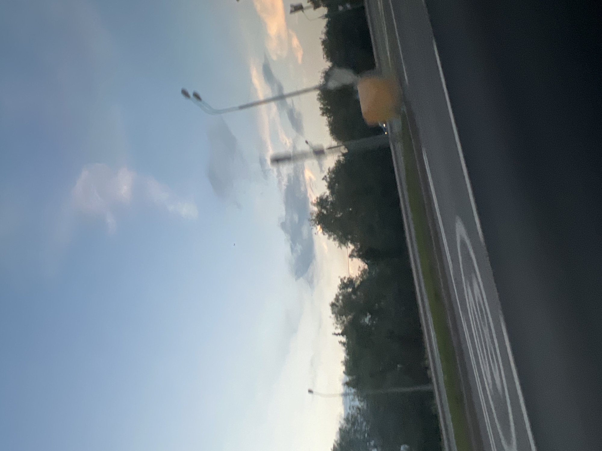 The image shows a view from inside a vehicle, looking out towards a street. The sky is visible, with light blue hues and some clouds. Streetlights are present along the road, which is bordered by trees. A speed limit sign is also visible next to the street. The location appears to be in a suburban or urban area.