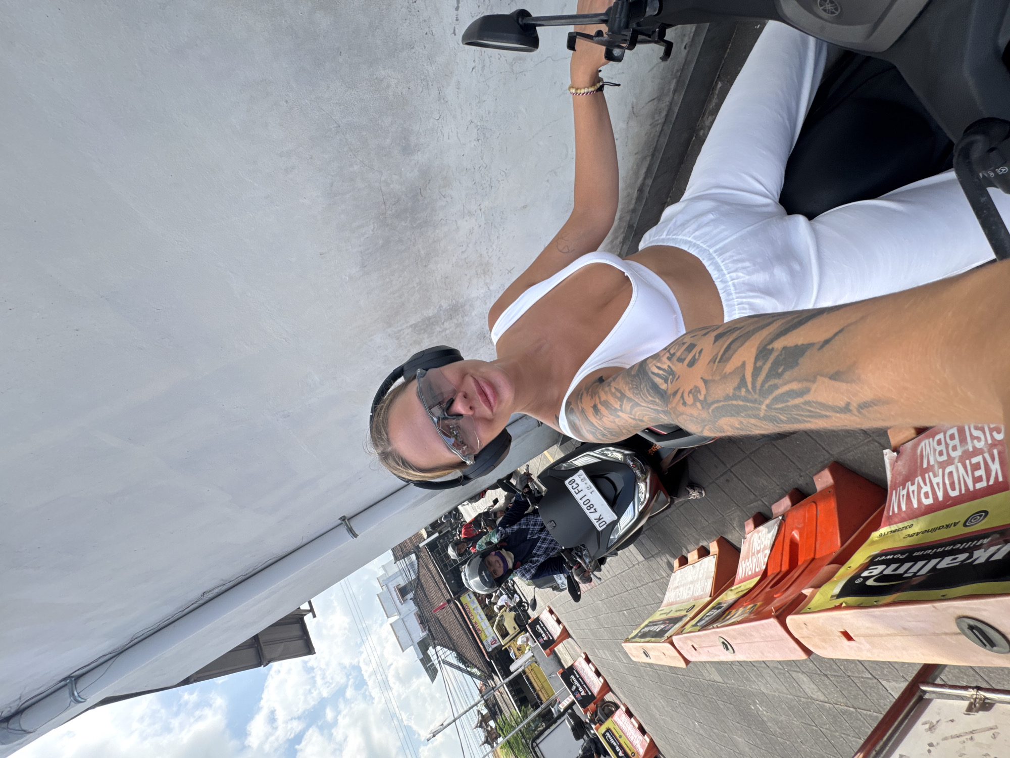 The image shows a woman in a white top and white pants wearing headphones and sunglasses taking a selfie while sitting on a motorbike. She has a tattoo on her left arm. Behind her, there are other people on motorbikes, orange and white plastic traffic barriers, and a sign with the words "KENDARAAN CISI BBM" and "Alkaline" on it. The motorbike in front of her has a license plate that reads "DK 4801 FC0." A partially visible sign reads “OPTIK CANGGU”. The location is North Kuta, Indonesia.