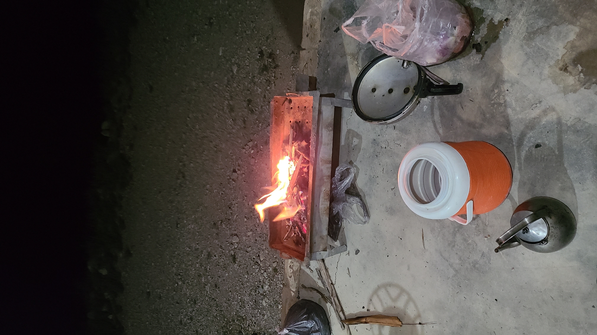 The image shows an outdoor scene, likely in Ziarat, Pakistan, given the presence of specific items. A small metal grill contains a fire. Next to the grill on the ground are a cooking pot lid, a plastic bag containing what appears to be food items, a large orange water cooler, and a metal kettle. A trash bag and some sticks lay at the bottom of the image. The surrounding environment appears to be a stone/gravel mixture that leads to darkness. The scene is lit artificially. No people are visible.