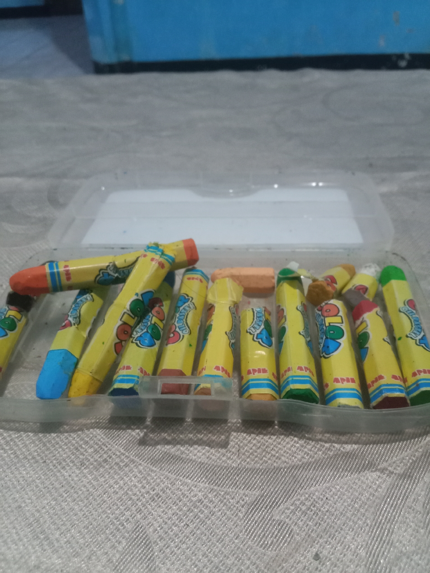 A clear plastic container holds a collection of brightly colored oil pastels. The pastels are mostly yellow with colorful labels that have blue, red, and green lettering. Some pastels have colored tips, including orange, blue, red, and green. The container is placed on a light-colored, textured surface, possibly a tablecloth or fabric. In the background, out of focus, are blurred blue shapes, suggesting furniture or a wall in the interior of a room. The lighting appears to be diffused, creating soft shadows. The image quality is slightly grainy. This scene is set in Kedungsari, Indonesia.