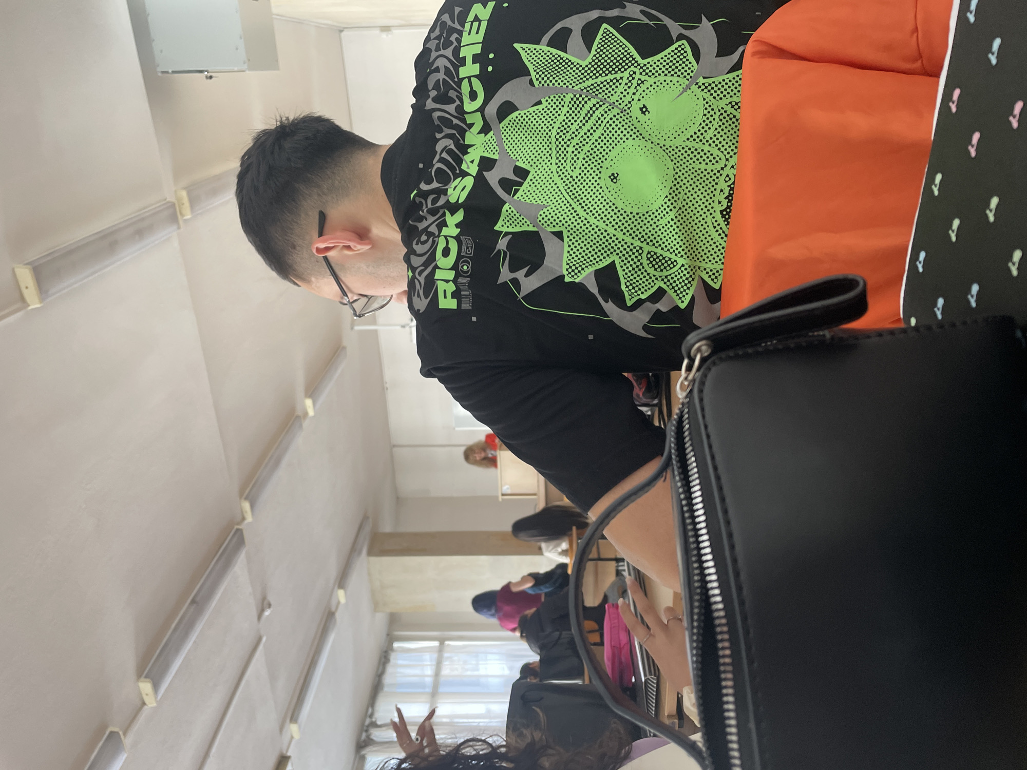 A student is in a classroom, likely in Varna, Bulgaria, based on the setting's typical characteristics of educational facilities. The student wears glasses and a black T-shirt with a large graphic of Rick Sanchez from the animated series "Rick and Morty," featuring green and gray colors. He sits in a classroom with other students, evident in the background with multiple individuals seated at desks. The room is lit by fluorescent lights. The photo is taken from a low angle and includes a black purse in the foreground. The setting appears to be a typical classroom environment.