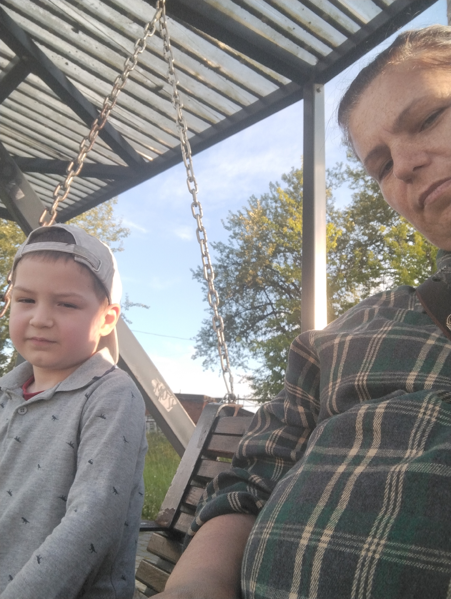 The image shows a selfie taken outdoors, likely on a swing set. In the foreground, a young boy wearing a grey baseball cap and a grey collared shirt with a pattern of small dark figures is seated on the left. To the right, an adult, presumably female, is also seated and partially visible, wearing a green and beige plaid shirt. The background shows trees, a blue sky with some clouds, and a structure that appears to be part of a playground or park, with metal chains and a wooden seat visible. The lighting suggests it is daytime. The location is stated to be the city of Kingisepp, Russia.