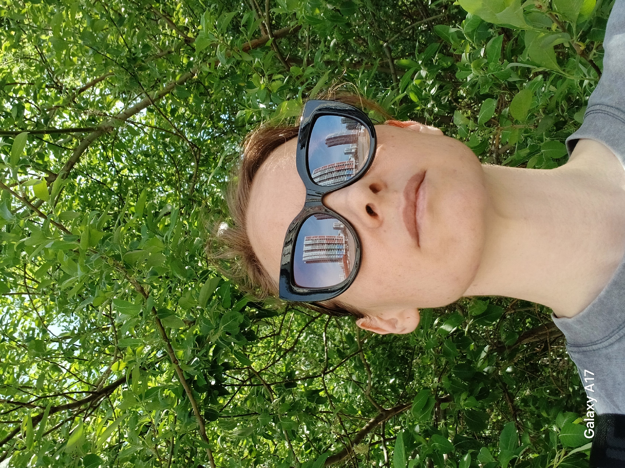 The image shows a person wearing black sunglasses, with a building reflected in the lenses. The background is filled with green foliage, likely from trees. The text "Galaxy A17" is visible in the lower right corner. The reflection in the sunglasses reveals a multi-story building with a distinctive architectural style, identifiable as a building located in Yekaterinburg, Russia.