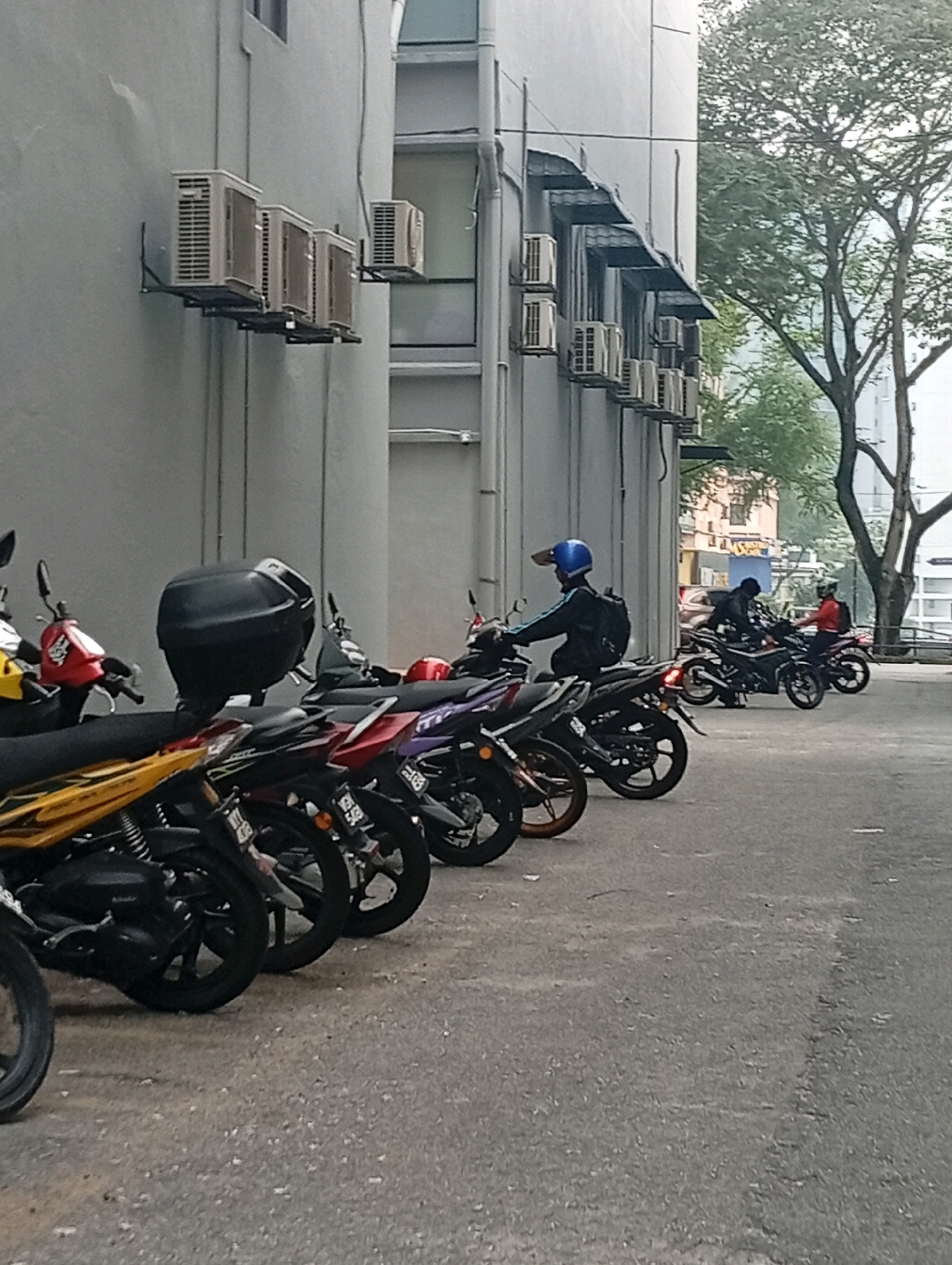 A row of motorcycles is parked in front of a light gray building, likely in Selayang Municipal Council, Malaysia. Air conditioning units are mounted on the exterior wall of the building. Several people are on or near the motorcycles. One person in a blue helmet is seated on a motorcycle in the foreground. Further down the row, two individuals in dark clothing are on separate motorcycles, and another person in a red jacket is also on a motorcycle. The ground is paved asphalt. Trees are visible in the background, suggesting an outdoor urban environment. The lighting indicates it is daytime.