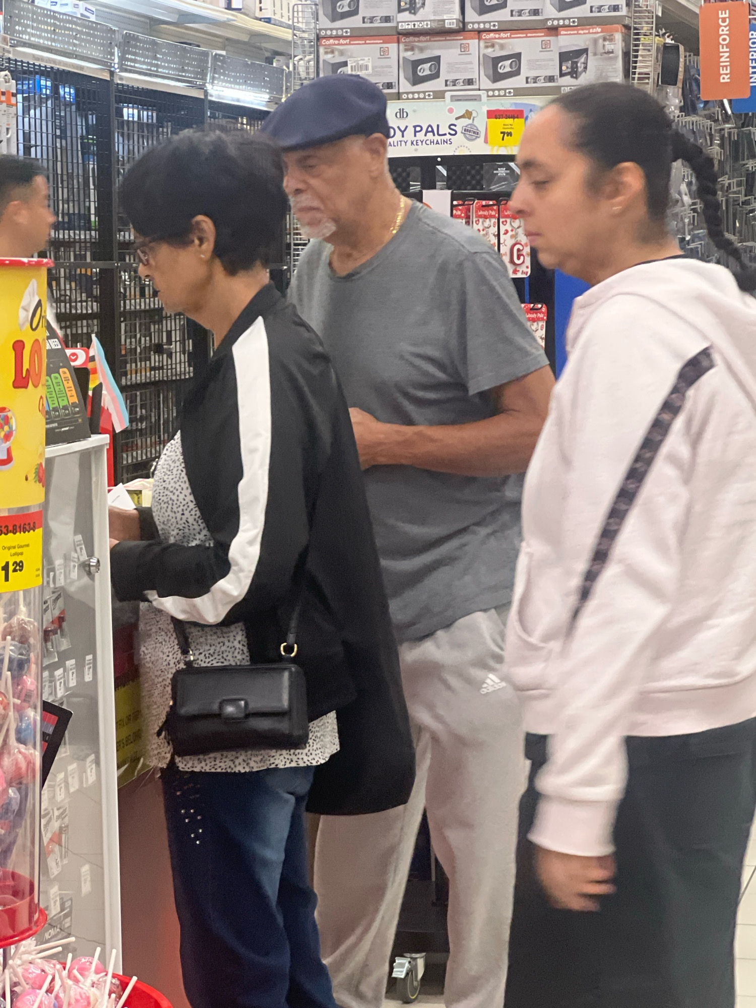 The image shows four individuals inside a store, likely a retail establishment such as a dollar store or discount retailer, given the merchandise displayed on shelves in the background. The scene appears to be an aisle with various products, including lollipops in a display case on the left.
On the far left, a person's profile is visible, looking to their right. Next to them, a woman with dark hair, glasses, and a black and white jacket is looking to her left. She is wearing a patterned top and dark jeans, and has a black purse with a gold clasp slung over her shoulder. She appears to be examining something at a shelf.
In the center, an older man with dark skin, a goatee, and a blue flat cap is wearing a grey t-shirt and grey sweatpants. He is looking forward and slightly to his right, appearing to be engaged in conversation or observation.
To the right of the man, a person with dark braided hair pulled back is wearing a light pink hooded sweatshirt. Their side profile is visible as they look to their left, toward the man.
The environment includes tall shelving units stocked with various items, some of which are packaged goods. A price tag that reads "$1.29" and indicates "Original Gourmet Lollipop" is visible on the left side of the frame. The lighting suggests an indoor commercial setting. The presence of a "REINFORCE" sign with orange lettering on the right suggests a department within the store. The image is taken at eye level. There is no specific indication in the image that places the scene in the city of Toronto, Canada.