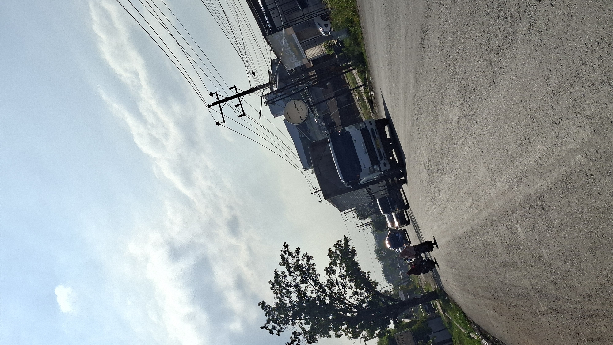 The image depicts a road scene in Keudebayu, Indonesia, under a partly cloudy sky. A paved road occupies the foreground, curving towards the right. On the left side of the road, a tall tree with dense foliage stands prominently. In the middle ground, traffic is visible, including a white truck with a dark cargo box, several cars, and two individuals on a motorcycle traveling in the same direction. The motorcycle riders are wearing headscarves. Beyond the road, a few buildings and structures are present, including a tall utility pole with multiple wires. The overall lighting suggests it is daytime, with sunlight casting shadows. The scene appears to be a moderately busy street in a semi-urban or rural area.