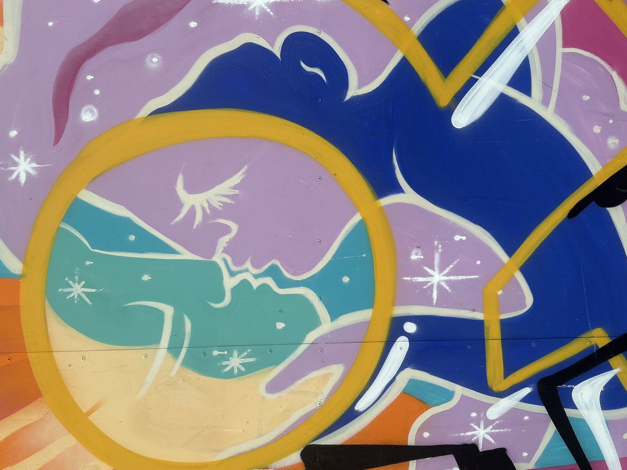 The image displays a vibrant graffiti mural on an outdoor wall, likely in Rafailovići, Montenegro. The artwork features stylized, abstract figures rendered in bold colors like blue, purple, teal, yellow, orange, and pink. A prominent circular yellow outline encloses two profile silhouettes facing each other, suggesting a kiss or intimate moment. These silhouettes are depicted in teal and lavender, with white outlines and accents. Scattered throughout the mural are white starburst shapes and small white dots, representing celestial elements. The background is a mix of pastel colors, including light yellow and lavender, with areas of solid blue. The graffiti also incorporates thick black and white lines, adding a graphic element to the composition. The surface of the wall appears slightly textured and shows some signs of wear, with visible rivets. The overall impression is one of cosmic romance or abstract celestial imagery.