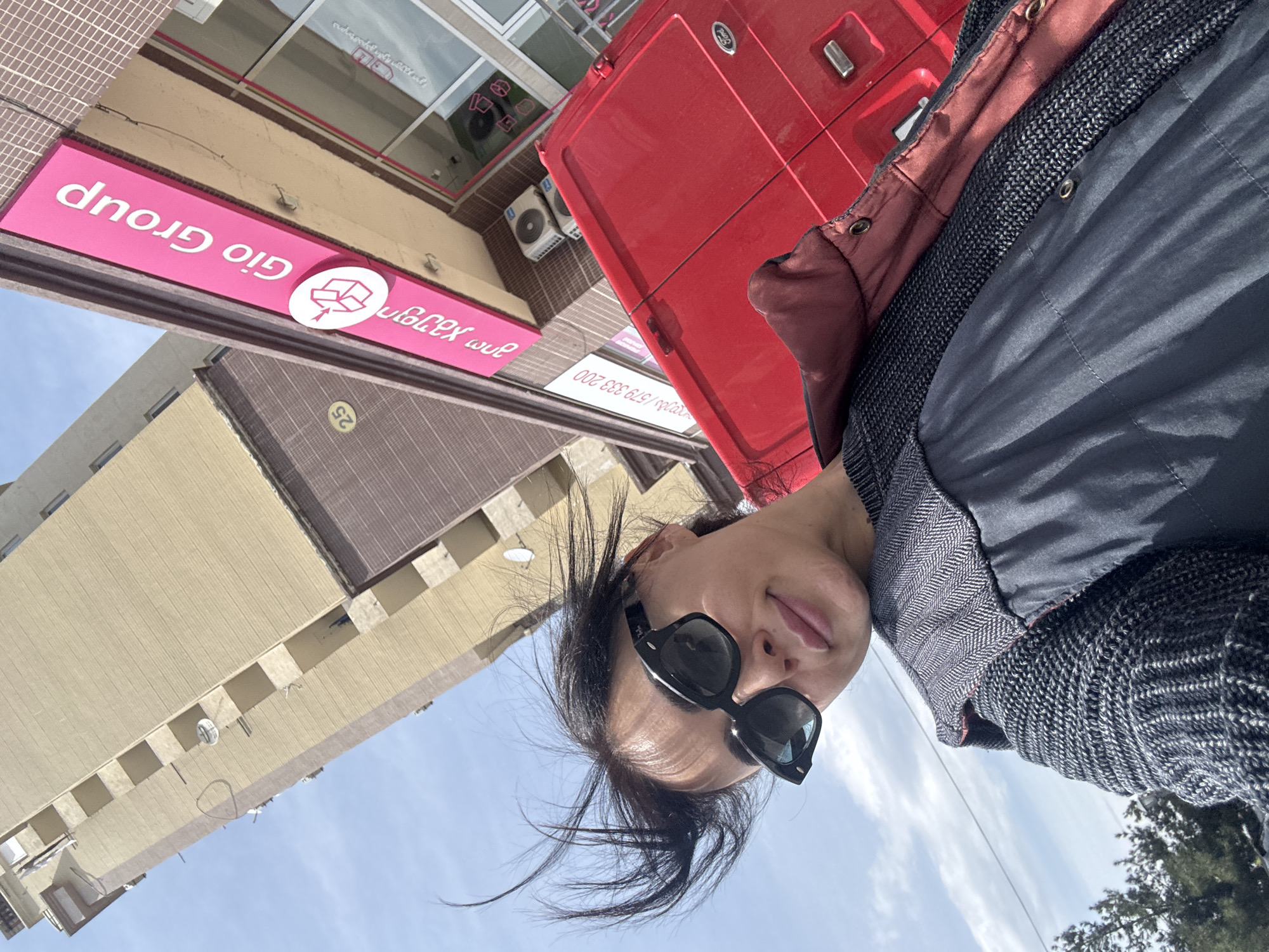 The image is a tilted, low-angle selfie taken outdoors on a bright, sunny day. In the foreground, a person, appearing female, is looking towards the camera with a neutral expression, wearing dark square-framed sunglasses. Her dark hair is slightly windblown around her face. She is dressed in a dark jacket with a reddish-brown lining, worn over a dark, textured knit sweater.
The background features urban elements, including buildings and a red vehicle. A prominent pink sign with white lettering is visible, displaying "Gio Group" in Latin script and "გიო ჯგუფი" (Gio Group) and "ჩეში გადამზიდი" (Cargo Carrier / Freight Forwarder) in Georgian script. Below the Georgian text, a phone number "579 333 200" is partially visible. The sign also includes a circular white logo with a stylized box. Another building, light beige in color, is visible, with a small yellow circular sign bearing the number "25".
A red Ford van or truck is positioned behind the person, partially visible, and its "Ford" logo is discernible. The sky above is clear blue with some scattered white clouds, indicating daytime conditions. The presence of Georgian script on the signboard aligns with the context of Tbilisi, Georgia.