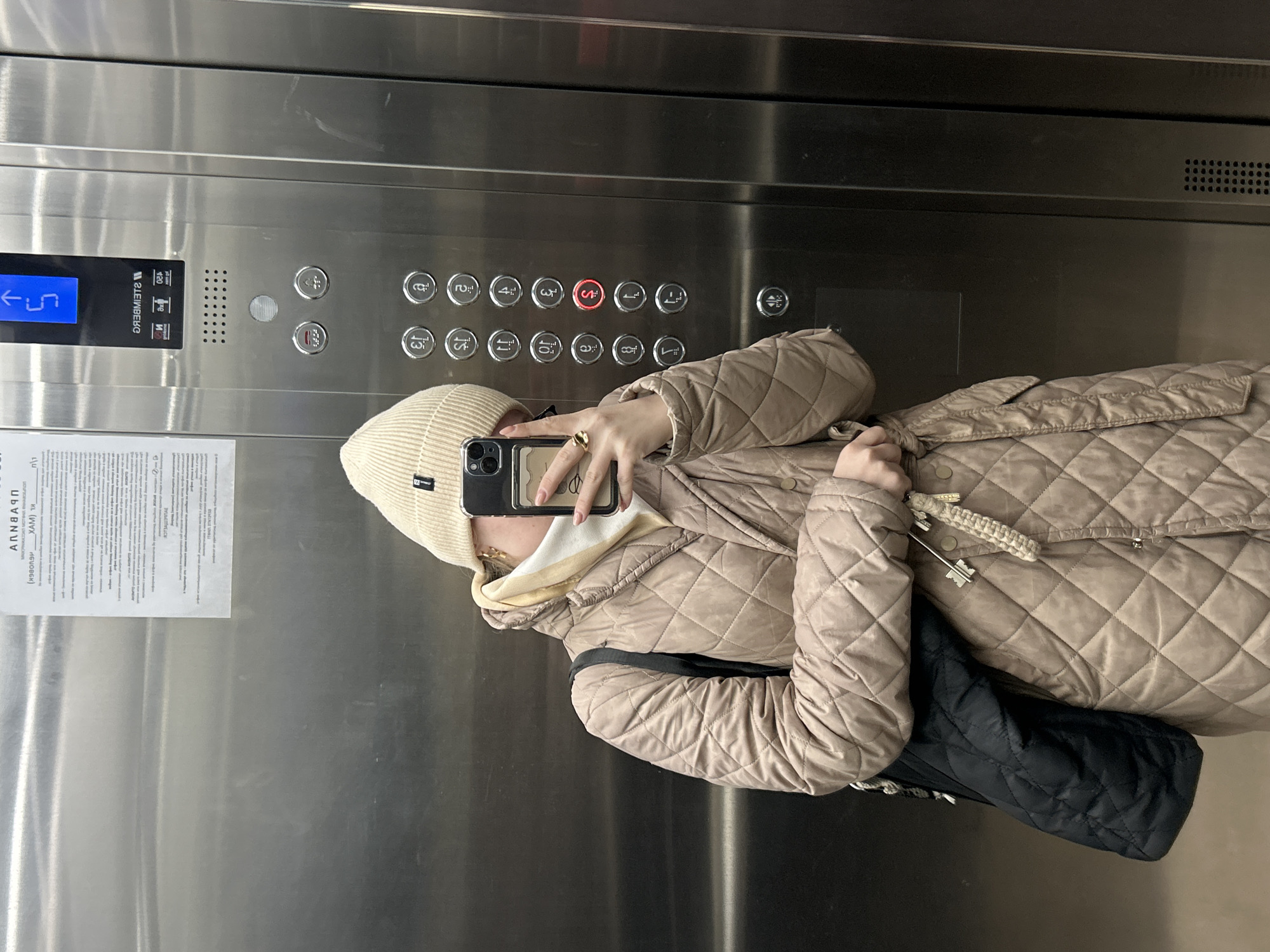 The image captures a person taking a mirror selfie inside an elevator. The person is wearing a light beige quilted jacket, a matching knit beanie, and a light-colored hooded top. They hold a smartphone to capture their reflection, which obscures most of their face. A golden ring is visible on their right hand, and a set of keys or a decorative charm hangs from their jacket. A black quilted bag is draped over their shoulder.
The elevator interior features polished stainless steel walls and a control panel. The digital display on the panel shows "← 5", indicating the current or target floor. The control panel contains a grid of circular buttons for various floors, ranging from "-1" (basement) to "15", including "G" (Ground) and "S" (Service/Special), with the "S" button illuminated in red. The brand name "ЛЕЙНБЕК" (LEINBEK) is prominently displayed on the panel. A white paper sign with partially legible Russian text, including "АППИА" (APPIA), is affixed to the elevator wall. The setting suggests a modern building, consistent with the Saint Petersburg context.