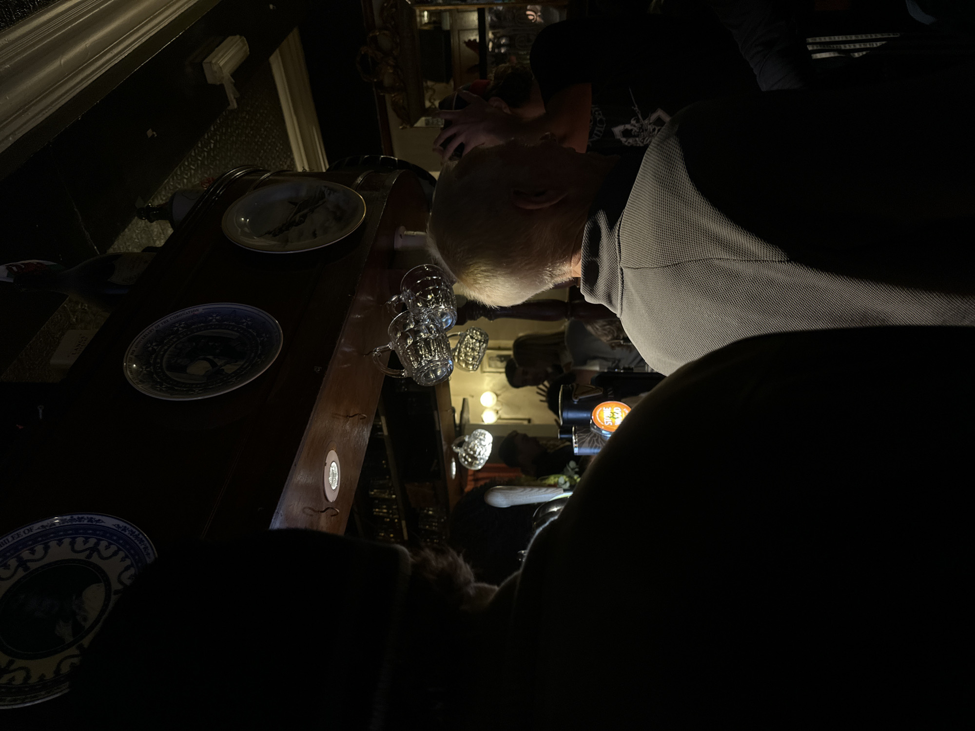 The image depicts an interior scene, possibly a pub or bar in London, United Kingdom. A man with short, light hair is leaning over a dark wooden bar surface. Several empty glass mugs and decorative plates are arranged on the bar. Behind the man, other patrons are visible in the dimly lit space, along with beer taps and light fixtures. The overall ambiance suggests an evening setting.