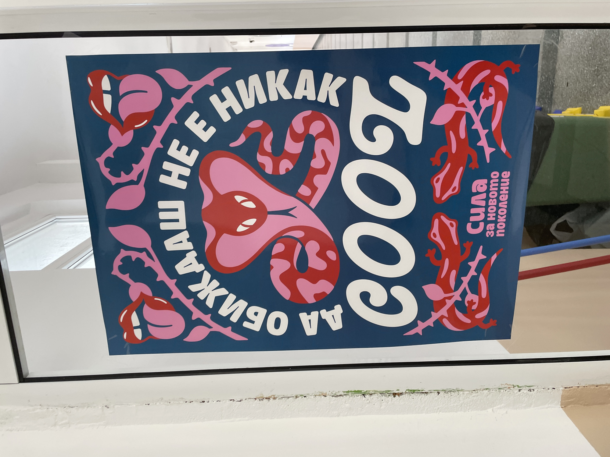 The image shows a poster stuck to a window. The poster is blue with pink and white designs. There is a snake in the center, surrounded by text in Bulgarian: "НЕ Е НИКАК" and "ДА ОБИЖАШ ЖИВОТ". The year "2002" is written vertically on the right side. There are stylized images of roses and lizards with thorns. A caption at the bottom right says "Сила за новото поколение" ("Strength for the new generation"). The window frame is white. Based on the text on the poster, it is likely located in Bulgaria. It is in Zlatograd.