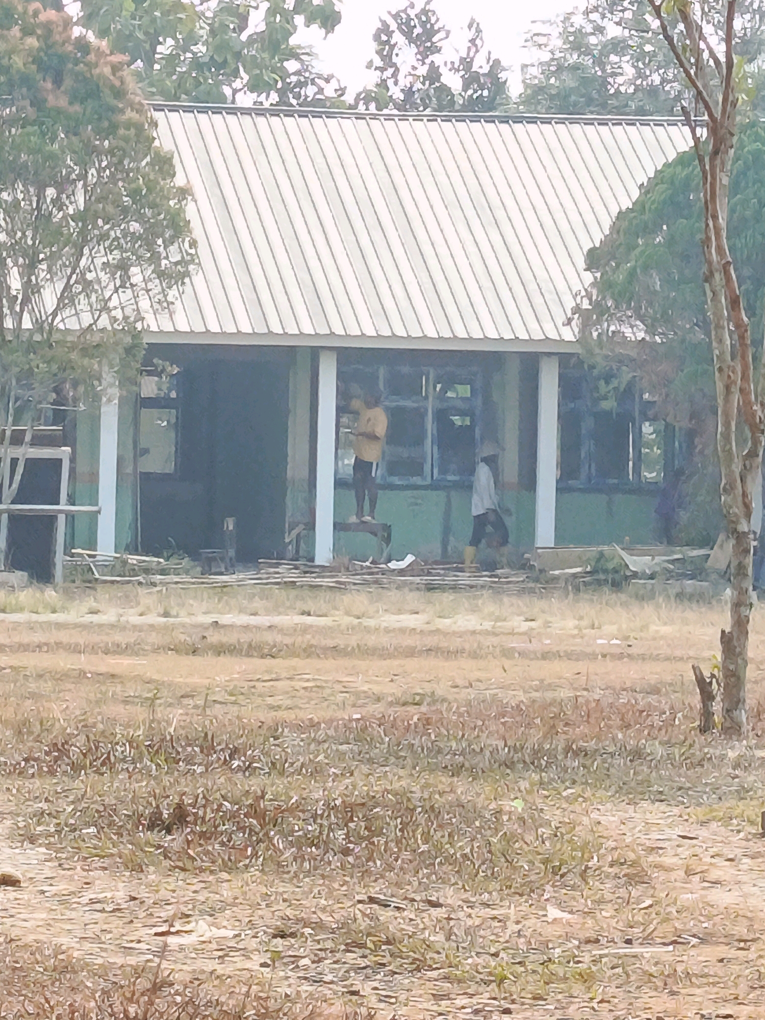 The image depicts a partially constructed or renovated building with a corrugated metal roof. Two individuals are present: one standing on a wooden plank in front of the building, looking at a phone, and another walking near the building entrance. The building's exterior is painted green, and it has several windows, some appearing to be boarded up. Scaffolding is visible on the left side of the building, and construction materials, such as wooden planks, are scattered on the ground in front of it. The environment appears to be an outdoor area with dry grass and some trees, suggesting a rural or semi-rural setting, potentially in Megang Sakti IV, Indonesia, given the context.