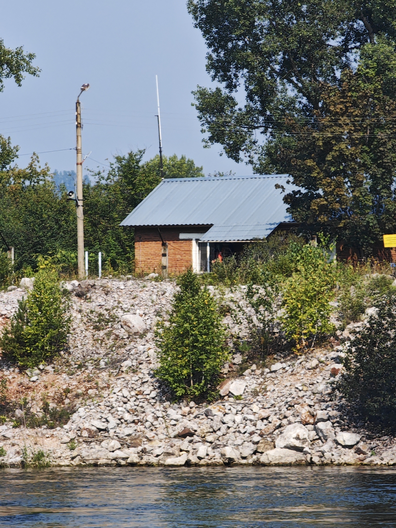 The image shows a small, brick building with a blue, corrugated metal roof, nestled on a rocky, overgrown slope. A large tree with dark green leaves dominates the right side of the frame, partially obscuring the building. In front of the building, there is a patch of rocky ground with sparse green vegetation. To the left of the building, a tall, light-colored pole with a light fixture and what appears to be a speaker or sensor is visible. The background consists of dense green trees and a clear blue sky. The foreground features the surface of a body of water, reflecting the sky and trees. The scene is an outdoor, natural environment with a human-made structure. It is not possible to determine the specific location within Есаулово, Russia, from the image alone. There are no people or identifiable actions visible.