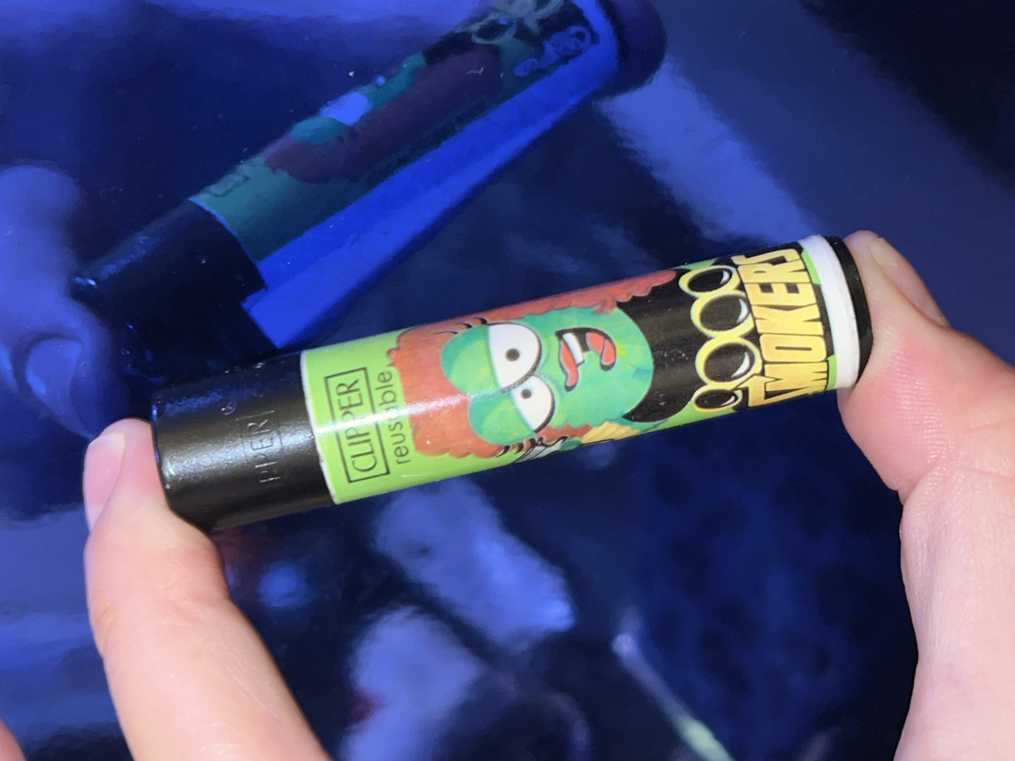 A person's hand holds a black, cylindrical object, likely a Clipper brand reusable lighter, against a highly reflective, dark blue surface. The object is wrapped with a vibrant green label featuring a cartoon character. This character has green skin, large white eyes, an open mouth with visible teeth, and scruffy orange or reddish hair.
Visible text on the green label clearly states "CLIPPER reusable". Further text spells out "SMOOOKERS", where the three "O"s are stylized as part of the cartoon character's eyes, and there is a small encircled "R" indicating a registered trademark.
The background is a smooth, glossy, dark blue surface that creates a strong, distorted reflection of the lighter and the hand. The lighting appears to be dim or casts a blue hue across the scene. The image focuses on the held object, with the person's fingers visible gripping its ends. No specific activity beyond holding the object is depicted. The overall impression is an close-up shot of the item. While the context given is Picassent, Spain, no visual elements within the image itself confirm this location.