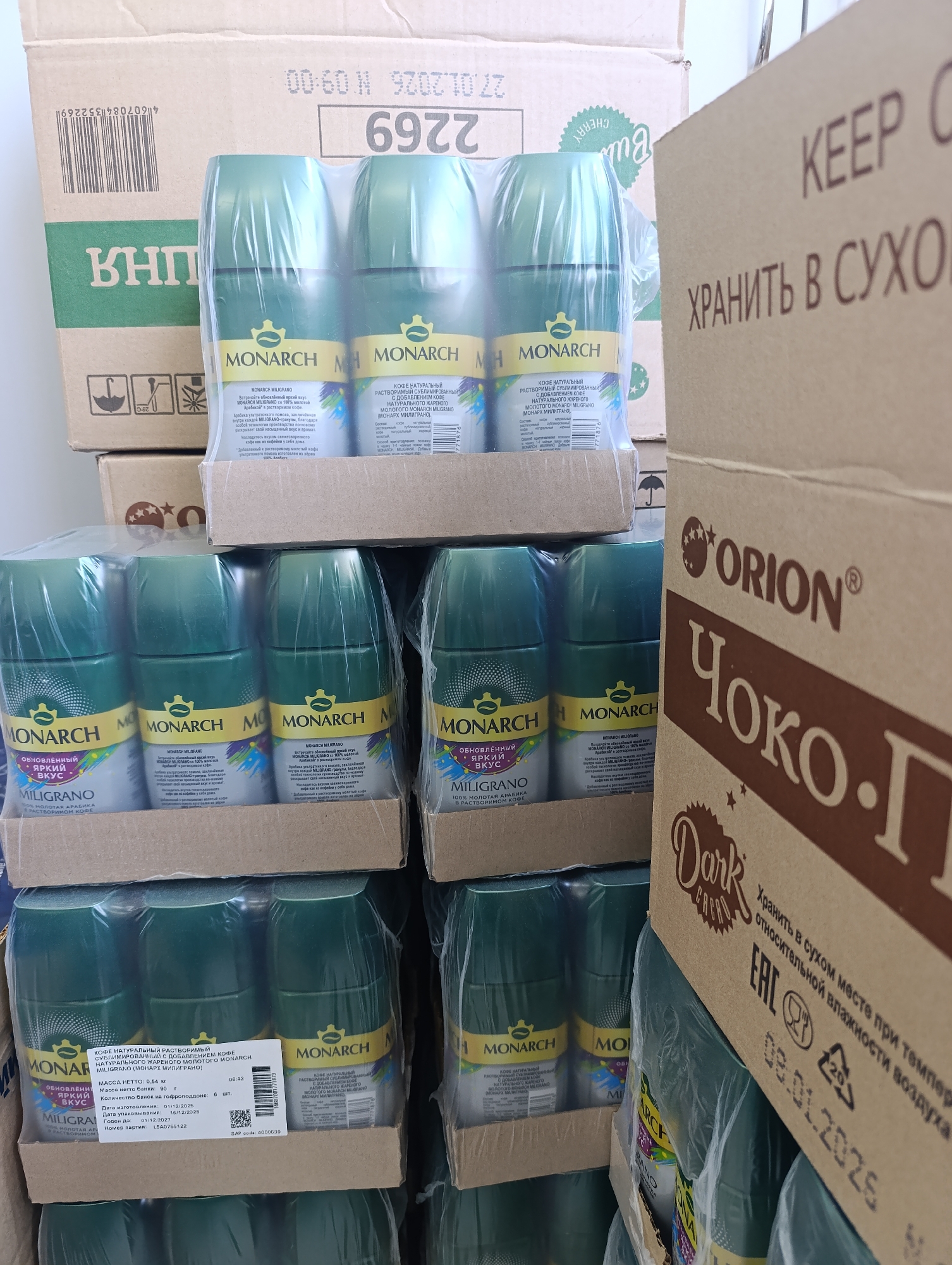 The image shows a stack of cardboard boxes, possibly in a storage area or warehouse. On the right side of the image is a box labeled "ORION Чоко-П". The other boxes appear to contain multiple packages of "Monarch Miligrano" instant coffee. The date "27.01.2026" and production code "6922" are visible on the top box. There's a close-up of a coffee package with the details: "Кофе Натуральный Растворимый Сублимированный с добавлением кофе Натурального Жареного Молотого Monarch Miligrano", along with Russian text. The production date is listed as 01/12/2025, and the expiration date is 01/12/2027. The image also features a barcode and SAP code 4000639.
Based on the Russian language and the presence of "Orion" products, the location is likely in a Russian-speaking region. While the image does not provide enough information to verify the precise location in the city of Chaikovsky, Russia, the labels on the boxes suggest that it is a place where goods like instant coffee and chocolate products are stored or sold.