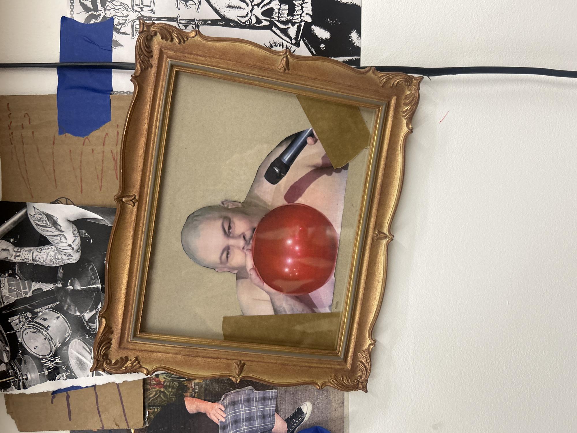 A close-up shot depicts a wall adorned with layered imagery. A gold-colored ornate picture frame is positioned prominently, displaying a collage or artwork of a bald man blowing up a red balloon while holding a microphone. The artwork is affixed to the frame with tape. The wall features additional images and materials, including black and white photographs of drums and people, scraps of cardboard with markings, and blue tape. A black cable runs horizontally across the top portion of the wall. The scene is captured indoors, likely in a studio or creative space. There are no people visible in the shot, just the image of the bald man inside the frame. The precise location in Los Angeles is impossible to determine from the provided image.