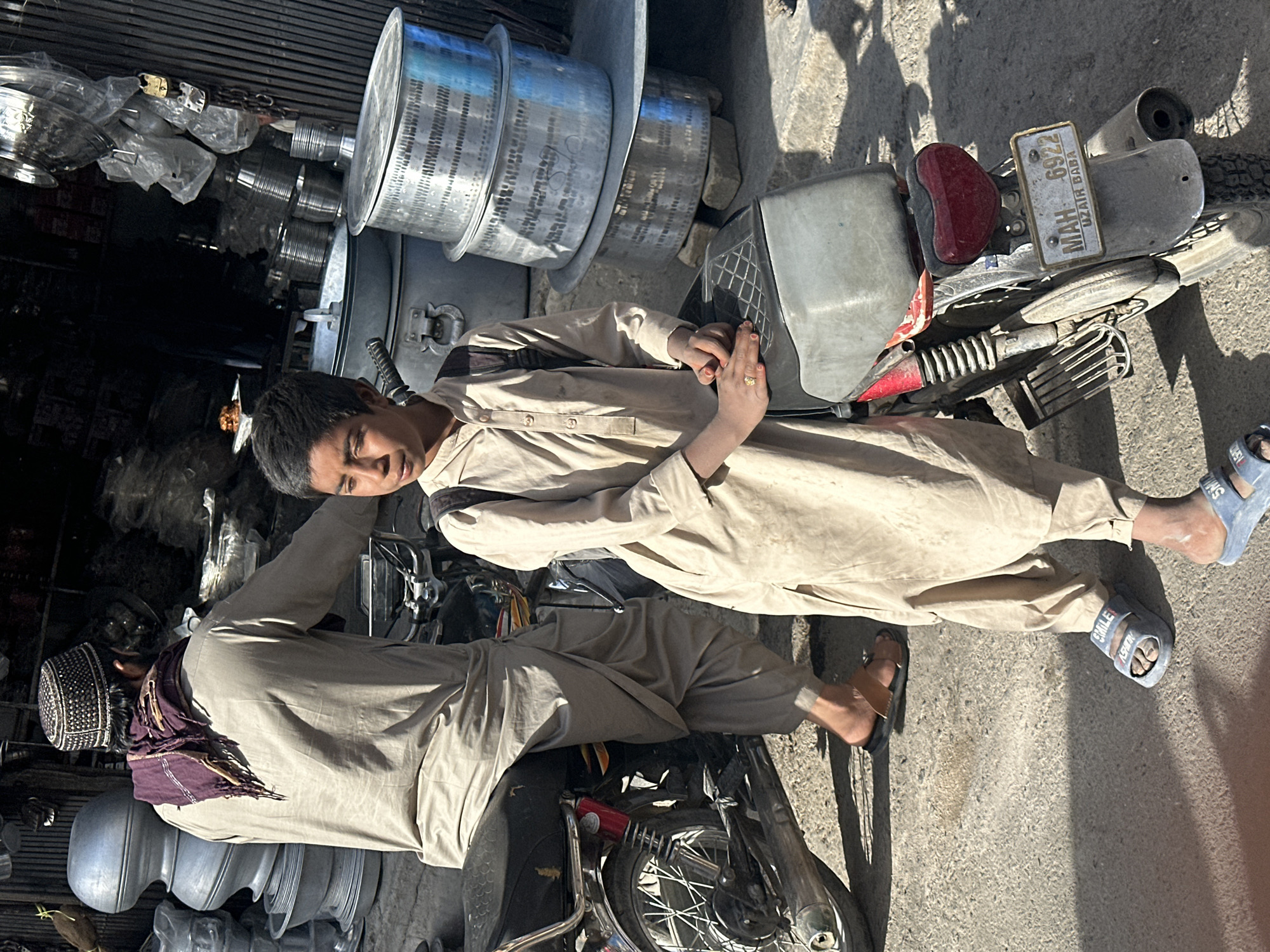 Two individuals are positioned on motorcycles, which are parked on a concrete or asphalt surface. The individual on the lower portion of the frame is wearing a dark-colored hat and a light-colored, possibly khaki, outfit. He is leaning towards the left side of the frame. The upper figure is clad in a similar colored outfit and is seated on the motorcycle, with his feet extending towards the right edge of the frame. He has light blue sandals on.
The motorcycles are of older models. The one partially visible at the bottom of the image features a red fender and a black tire. The motorcycle on which the seated individual is positioned has a license plate with the text "MAH 6922 UZAIR BABA".
In the background, to the left, is an array of shiny, metallic cookware and containers, stacked and arranged for sale, suggesting a commercial setting. The environment looks urban, possibly a street-side market or shop in an area known for selling kitchenware.
The image captures a casual moment with individuals interacting with motorcycles in a commercial area, likely during daylight hours. Location is Unknown, Unknown.