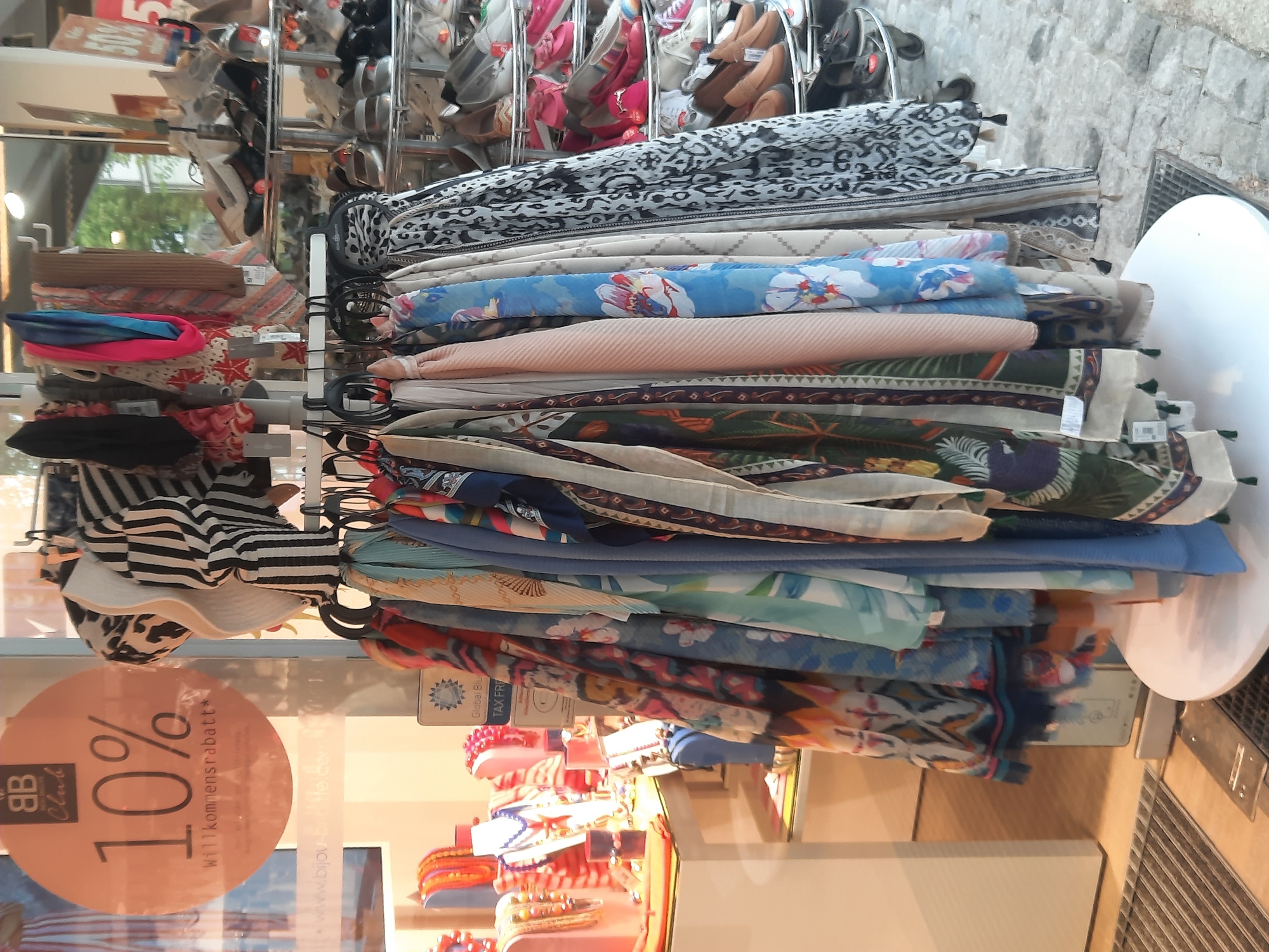 In Rosenheim, Germany, an outdoor display of scarves is set up in front of a storefront. A circular sign with "BB Club" and "10% Willkommensrabatt" is prominently displayed, suggesting a welcome discount for new customers. The scarves are arranged on a multi-tiered rack, showcasing a variety of patterns and colors, including floral prints, geometric designs, and solid hues. Behind the scarves, racks of shoes are visible, indicating that the store likely sells both accessories and footwear. The scene appears to be on a cobblestone street, with a metal grate in the foreground.