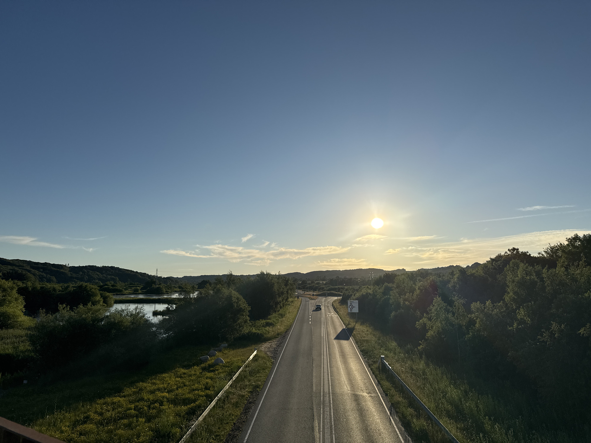 The image captures a scenic view of a road leading towards a distant horizon, bathed in the warm glow of a setting sun. The road, asphalt and marked with white lines, is flanked by lush green vegetation, including trees and grassy fields dotted with yellow wildflowers. To the left, a tranquil body of water reflects the sky. In the distance, rolling hills create a picturesque backdrop. A single car is visible on the road, heading away from the viewer. A traffic sign indicating a roundabout is present on the right side of the road. The sky is a clear blue, with a few wispy clouds scattered across it. The image appears to be taken from an elevated perspective, possibly a bridge or an overlook, looking down onto the road and the surrounding landscape. The location is likely the Vejle municipality in Denmark, given the presence of similar natural landscapes and road infrastructure often found in the region.