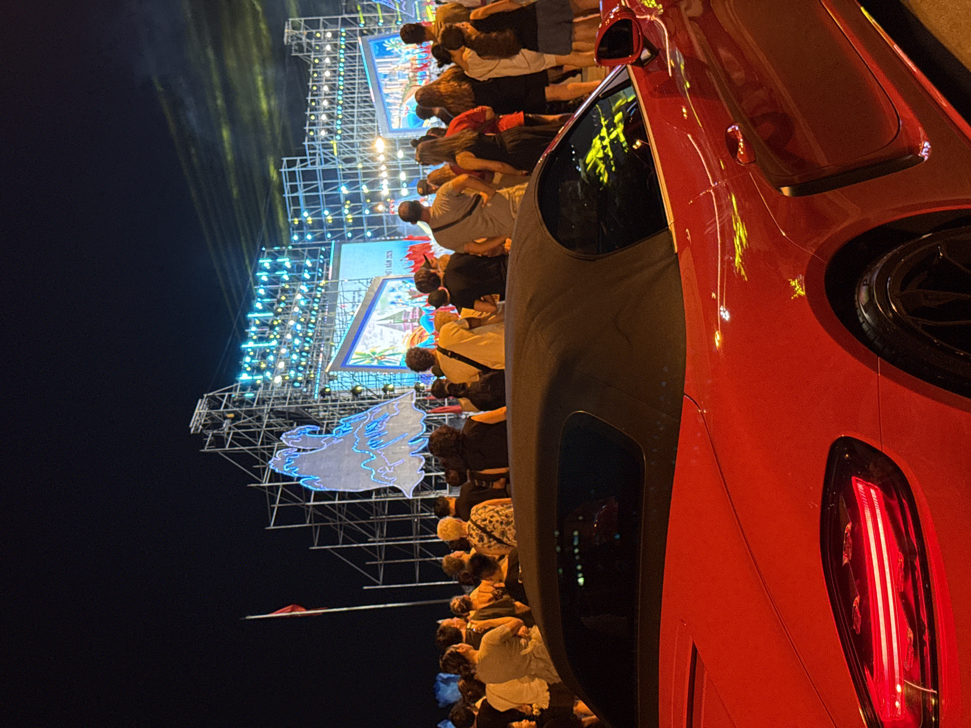 This image captures a night scene in Nha Trang, Vietnam, likely during a festival or event. In the foreground, a bright red convertible sports car is parked, its sleek design illuminated by ambient lights. The car's red taillight is prominently visible, and its roof is down. Behind the car, a crowd of people stands facing a large stage or elaborate light installation. The stage is adorned with numerous bright lights, giving it a festive and energetic appearance. In the background, there's a tall structure, possibly scaffolding or a temporary stage support, covered in lights. A red flag on a flagpole is visible to the left of the stage. The overall atmosphere suggests an outdoor public gathering with entertainment.