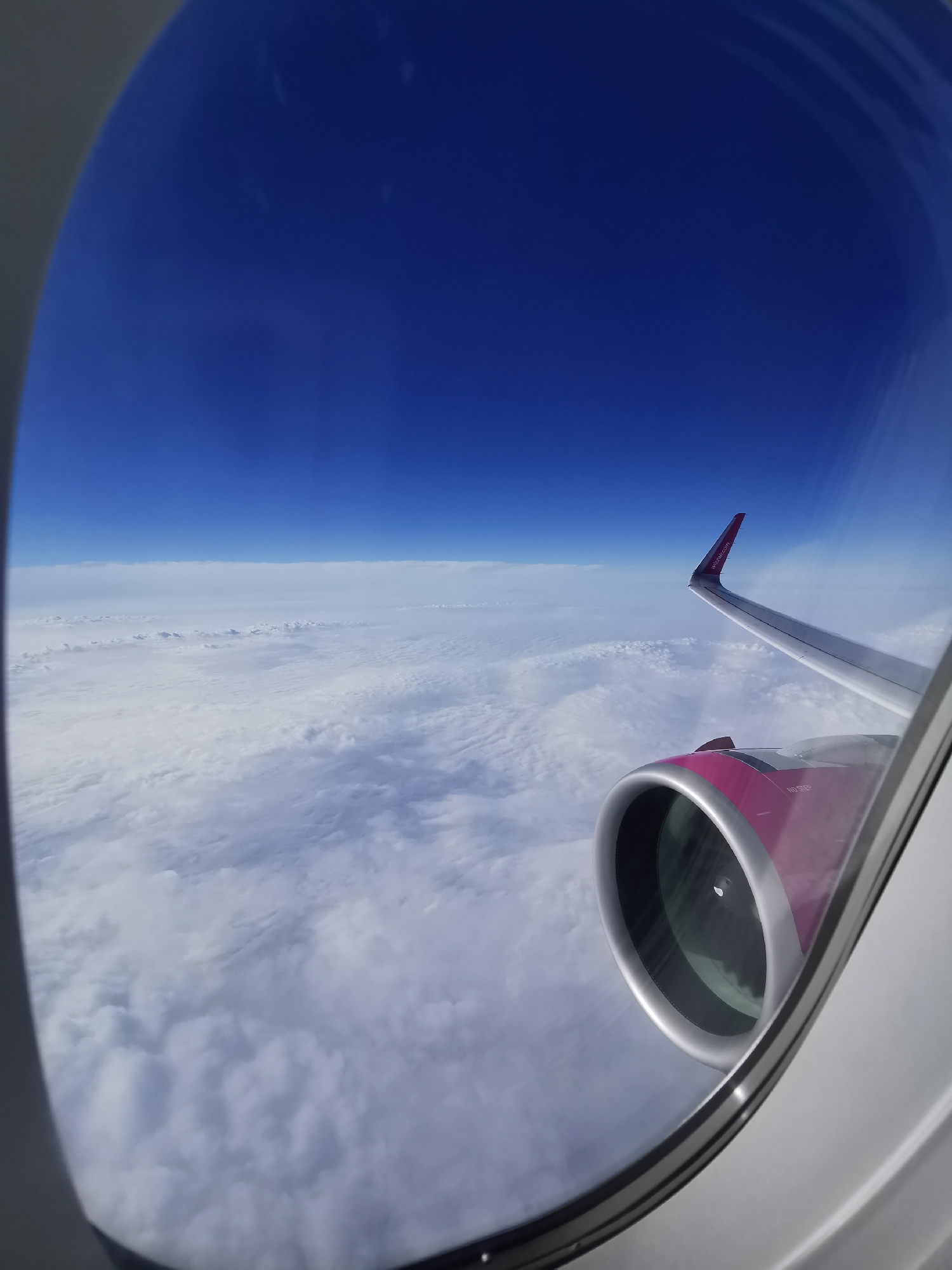 View from an airplane window showing a wing and engine, with clouds and blue sky visible. The tail wing is branded with "Wizzair.com," but the exact location in Varna, Bulgaria is not discernible.