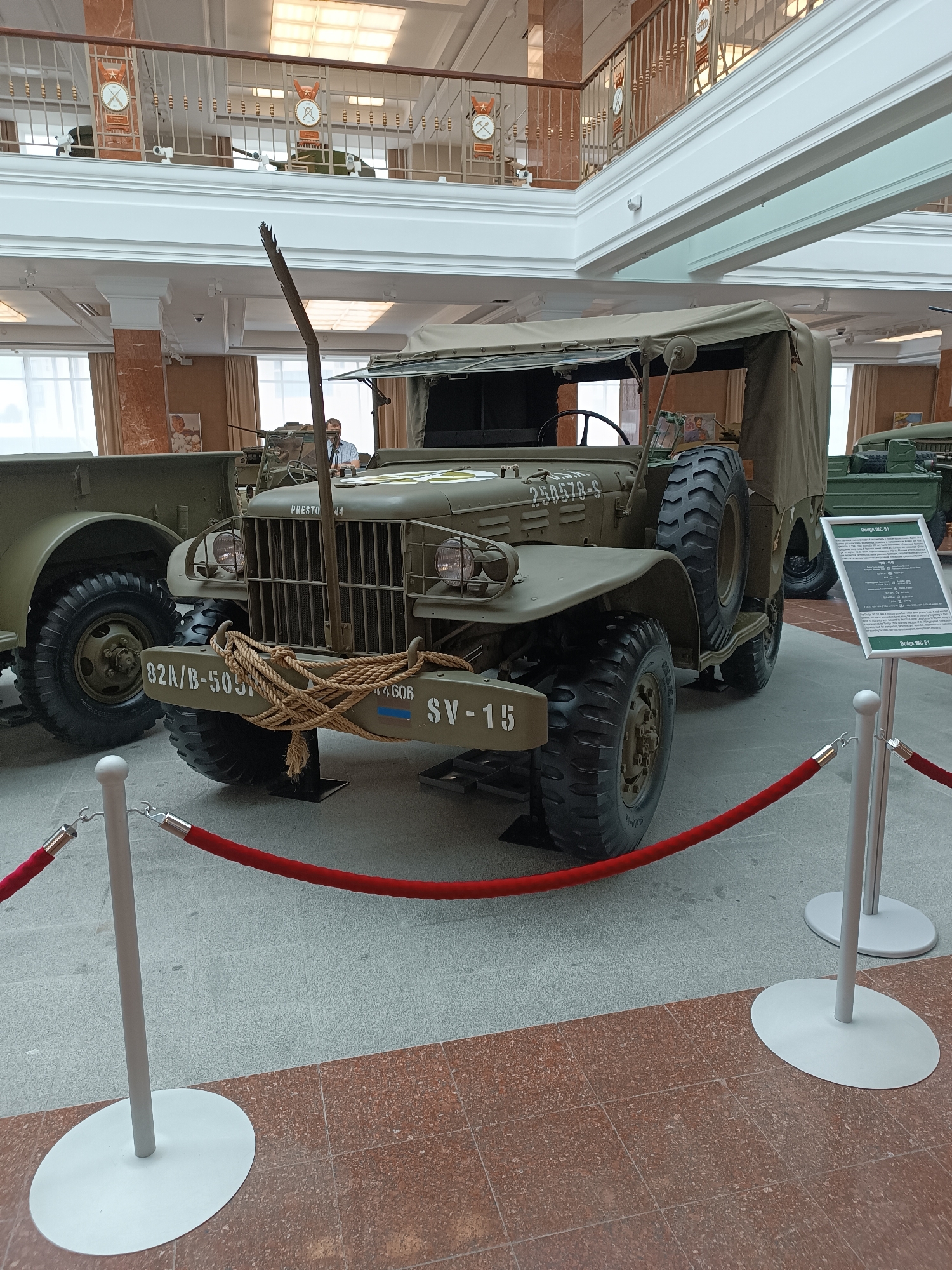 This is a medium shot, eye-level photograph of a vintage olive-drab military truck, likely a Dodge WC-51, on display inside a museum. The truck is the central focus, positioned at an angle to showcase its front and right side. It has a canvas top, large rugged tires, and a spare tire mounted on the right rear fender. A thick rope is wrapped around the front bumper. The vehicle appears to be in excellent condition, suggesting it is well-maintained for exhibition.
The setting is a museum hall with a polished floor, a mix of grey and red-brown tiles, and high ceilings with ornate white molding and recessed lighting. In the background, through large windows, daylight can be seen. A red velvet rope barrier with silver stanchions is in the foreground, indicating that the truck is roped off from visitors. To the right of the truck, a sign provides information about the vehicle, identifying it as a Dodge WC-51. Behind the truck, another military vehicle is partially visible. On an upper level of the museum, visible in the background, there are railings and displays that hint at further exhibits. There is no discernible activity or event taking place beyond the static display of the vehicles. The lighting suggests it is daytime. A few people are visible in the far background, but they are not prominent.
