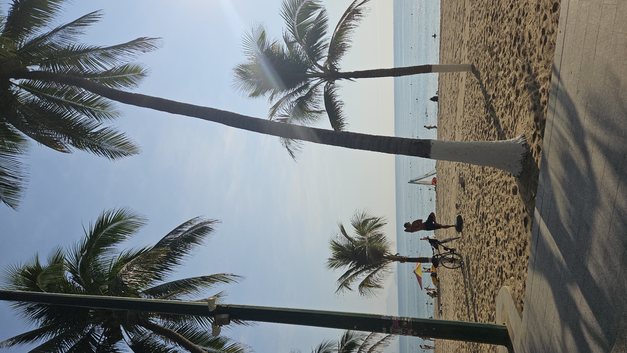 Here's a forensic description of the image:
**Location:** Coastal area in Nha Trang, Vietnam, likely a beach promenade.
**Environment:** Sunny day with a clear blue sky and a calm ocean. Sandy beach with some wave action visible in the distance.
**Objects:**
* Several tall palm trees with painted white trunks.
* A paved walkway alongside the beach.
* Bicycle parked on the sand.
* A few distant windsurfing sails.
* Street light poles.
**People:**
* A man is lying on the beach with a towel or mat, seemingly relaxing.
* A few distant figures are visible on the beach and in the water.
**Action/Interactions:**
* The man is in a reclined position on the beach.
* Other people are swimming and walking along the beach.
* The scene suggests a relaxed and typical beach atmosphere.